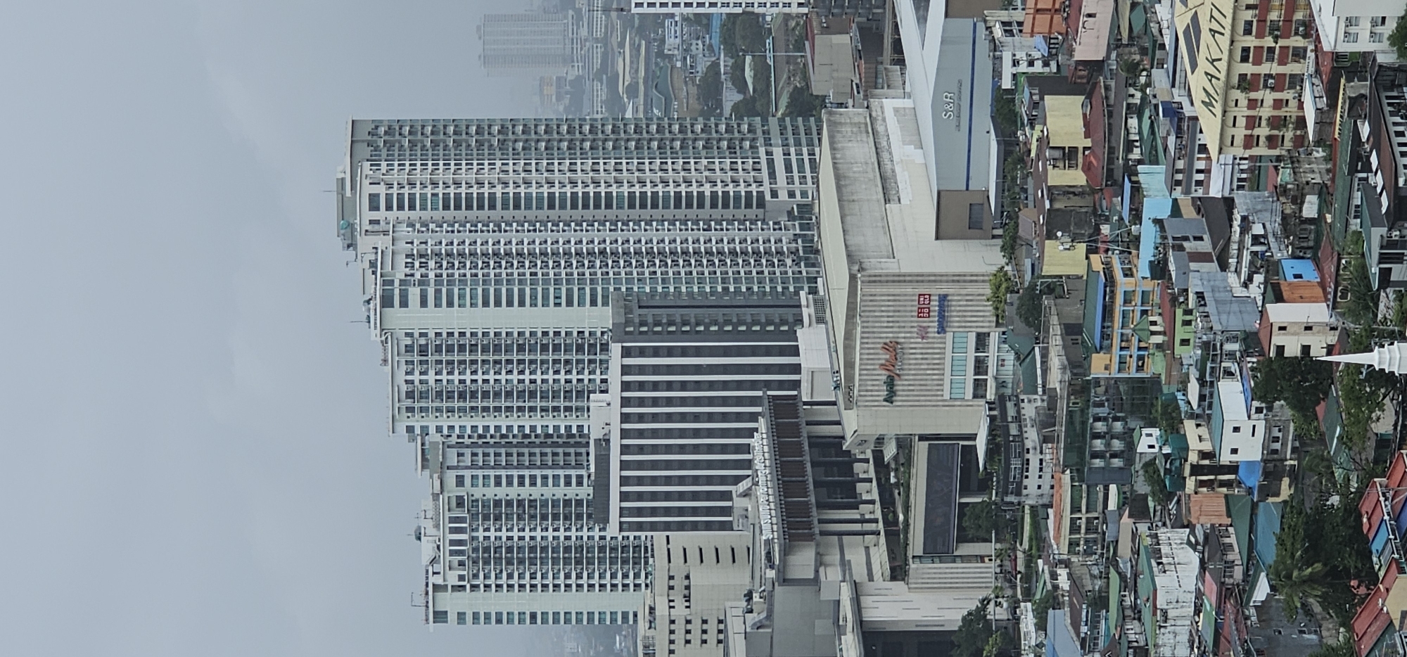 This is a vertical, eye-level, wide shot of a dense urban landscape in Makati, Philippines, dominated by modern high-rise buildings and older, lower-rise structures. The sky is overcast and a muted gray, suggesting a cloudy or hazy day.
In the foreground and mid-ground, several tall, slender residential towers with numerous balconies and windows rise prominently. They have a consistent, modern architectural style, primarily in light colors like white and pale green. Between these towers are mid-rise buildings, including a multi-story building with vertical dark stripes and a large, flat-roofed complex labeled "Ayala Malls" and "S&R". Adjacent to the mall complex, a large, cream-colored building has the word "MAKATI" emblazoned across its top.
Below the modern complexes, the scene transitions to a dense network of older, lower-rise buildings. These structures are varied in color and architectural style, with many featuring colorful rooftops (green, red, blue) and visible air conditioning units. The density of these buildings suggests a mixed-use urban environment with residential and commercial activity.
In the far distance, more high-rise buildings can be seen, some appearing lighter and possibly further away due to atmospheric haze. The overall impression is one of a bustling, rapidly developing city with distinct layers of development, from the contemporary skyscrapers to the more established, older districts. There are no visible people or specific actions depicted in the image.