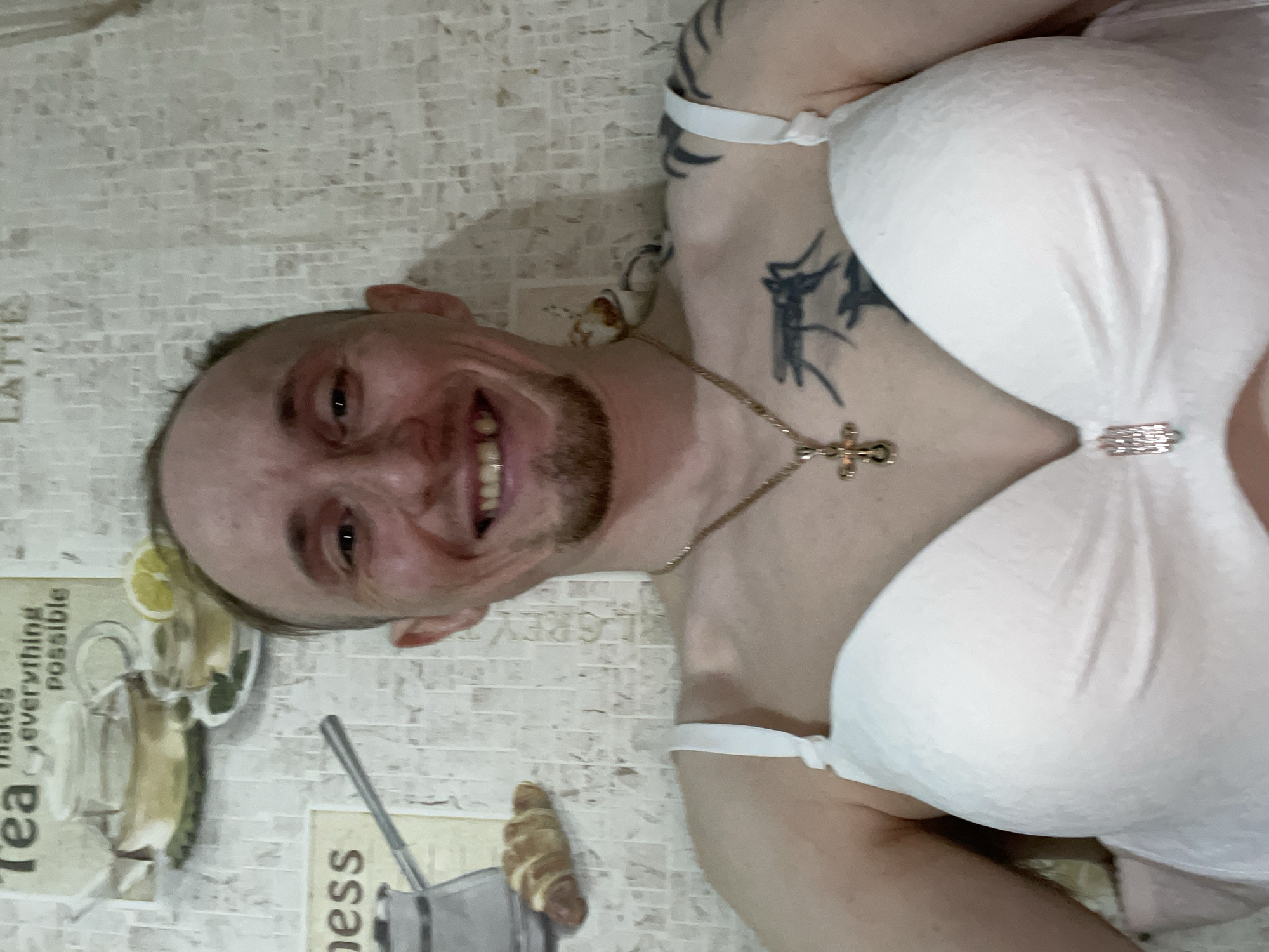 A man is lying down wearing a white bra. He has a gold chain with a cross pendant around his neck and tattoos on his chest and arm. The background features a wall with a design of tea, a lemon, and kitchenware, possibly in a location with a breakfast or kitchen theme. It is unknown if the city of Devyatovo, Russia, is specifically relevant to the scene depicted.