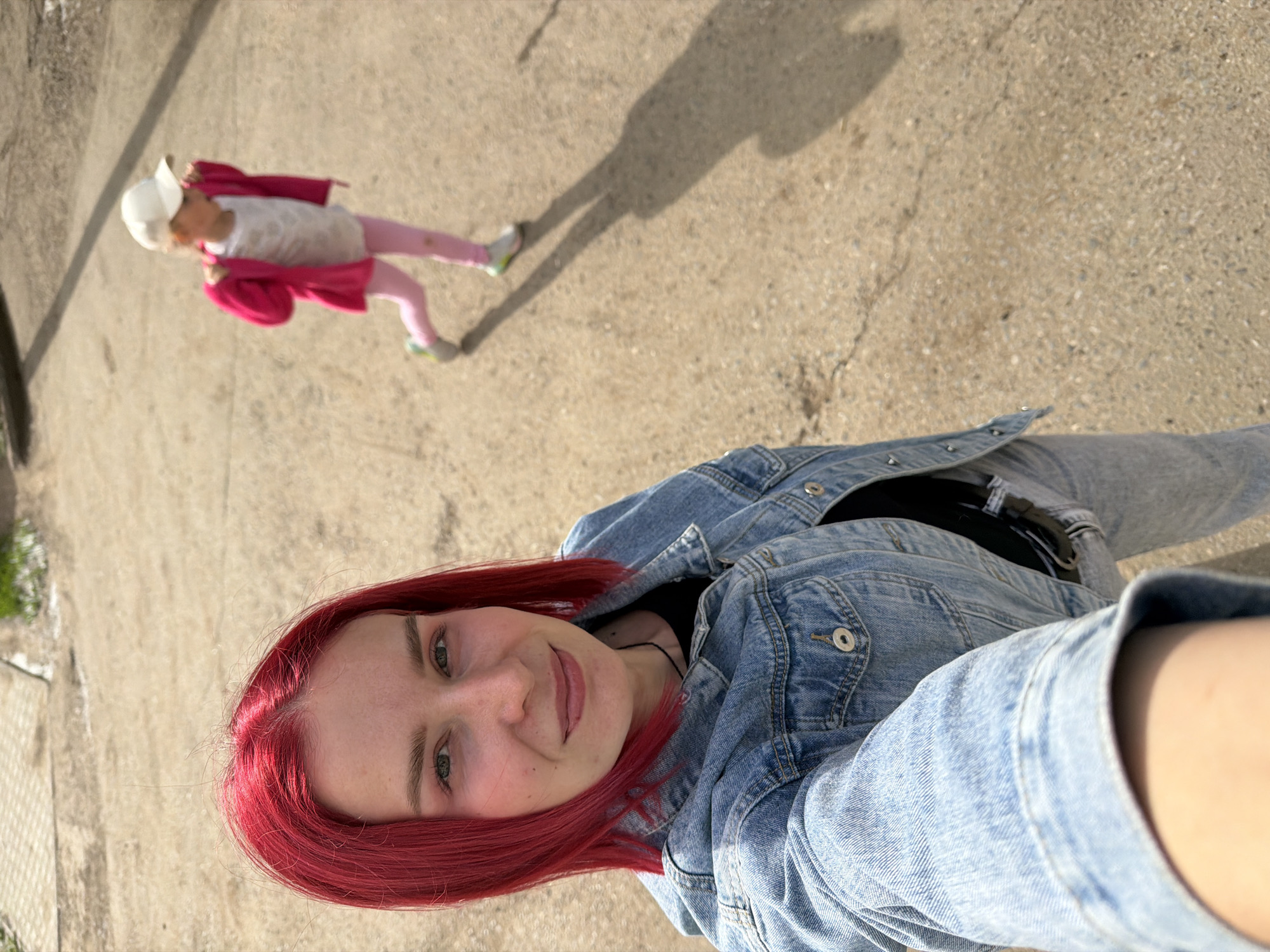 The image is a bright, sunny outdoor selfie featuring a woman in the foreground and a child in the background. The woman, who has vibrant red hair and light-colored eyes, is smiling slightly and looking directly at the camera. She is wearing a light blue denim jacket over a black top and light-wash denim jeans. Her arm is visible in the lower right, holding the camera for the selfie.
In the blurred background, a young child, likely a girl, is walking away from the camera. She is dressed in a white baseball cap, a bright pink long-sleeved top or jacket, pink pants, and light-colored sneakers with green detailing. A distinct shadow of the child stretches across the ground behind her, indicating strong sunlight.
The setting is an outdoor paved area, likely a concrete street, sidewalk, or path, showing some cracks and texture. A small strip of green vegetation is visible along the left edge. The presence of clear shadows suggests it is daytime, possibly late morning or afternoon, with clear weather. The overall mood of the image is casual and cheerful. No visible text is present. The scene is situated in an urban or suburban outdoor environment in Chaikovsky, Russia.