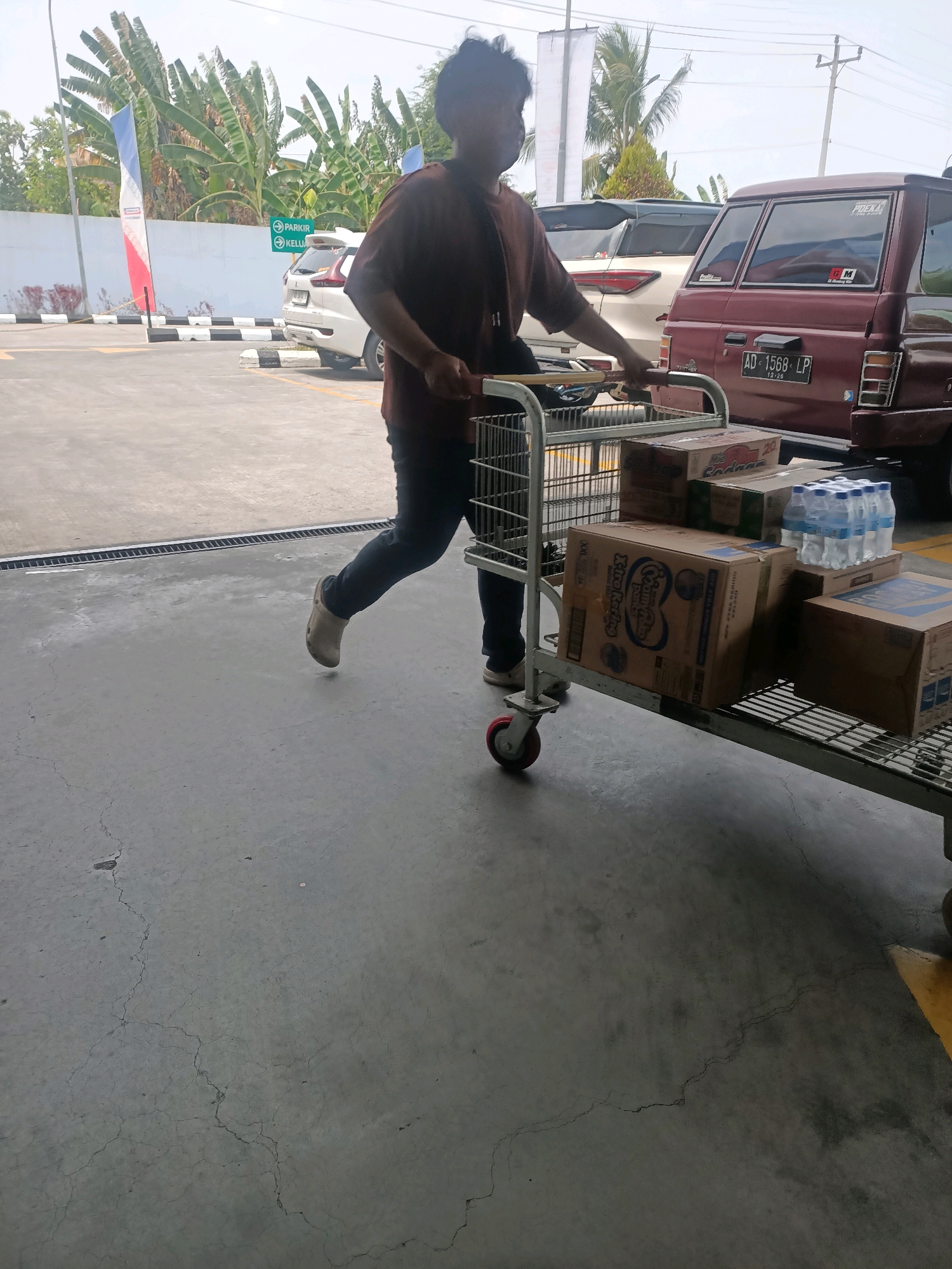 In Jaten, Indonesia, a person is seen pushing a shopping cart filled with various items, including boxes of goods and bottles of water. The shopping cart is made of metal with red wheels and is positioned on a cracked concrete surface. The person is wearing a brown shirt and dark pants, and they appear to be in motion. In the background, several cars are parked, including a maroon vehicle with a license plate that reads "AD 1568 LP". Other vehicles and structures, such as a white wall and signage, are also visible. Banana trees and a flag are present in the background, suggesting an outdoor location, possibly a parking lot or an area adjacent to a commercial establishment. The sky is overcast.