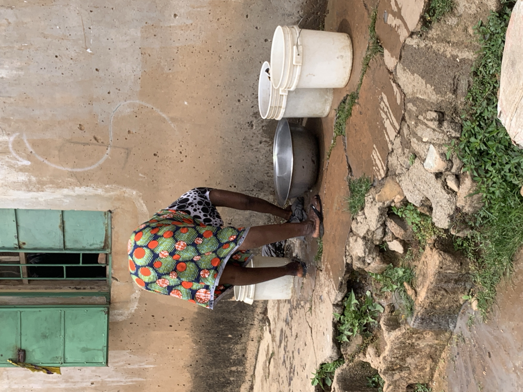 A person stands outside a tan-colored building with a green window in Jos, Nigeria. The individual is wearing a skirt with orange and blue polka dots over a green background and is bent over a collection of buckets and a silver bowl. Water appears to be flowing on the ground near a collection of rocks and greenery. Three white buckets are stacked near the bowl. The ground is concrete and cracked. The window is partially open.