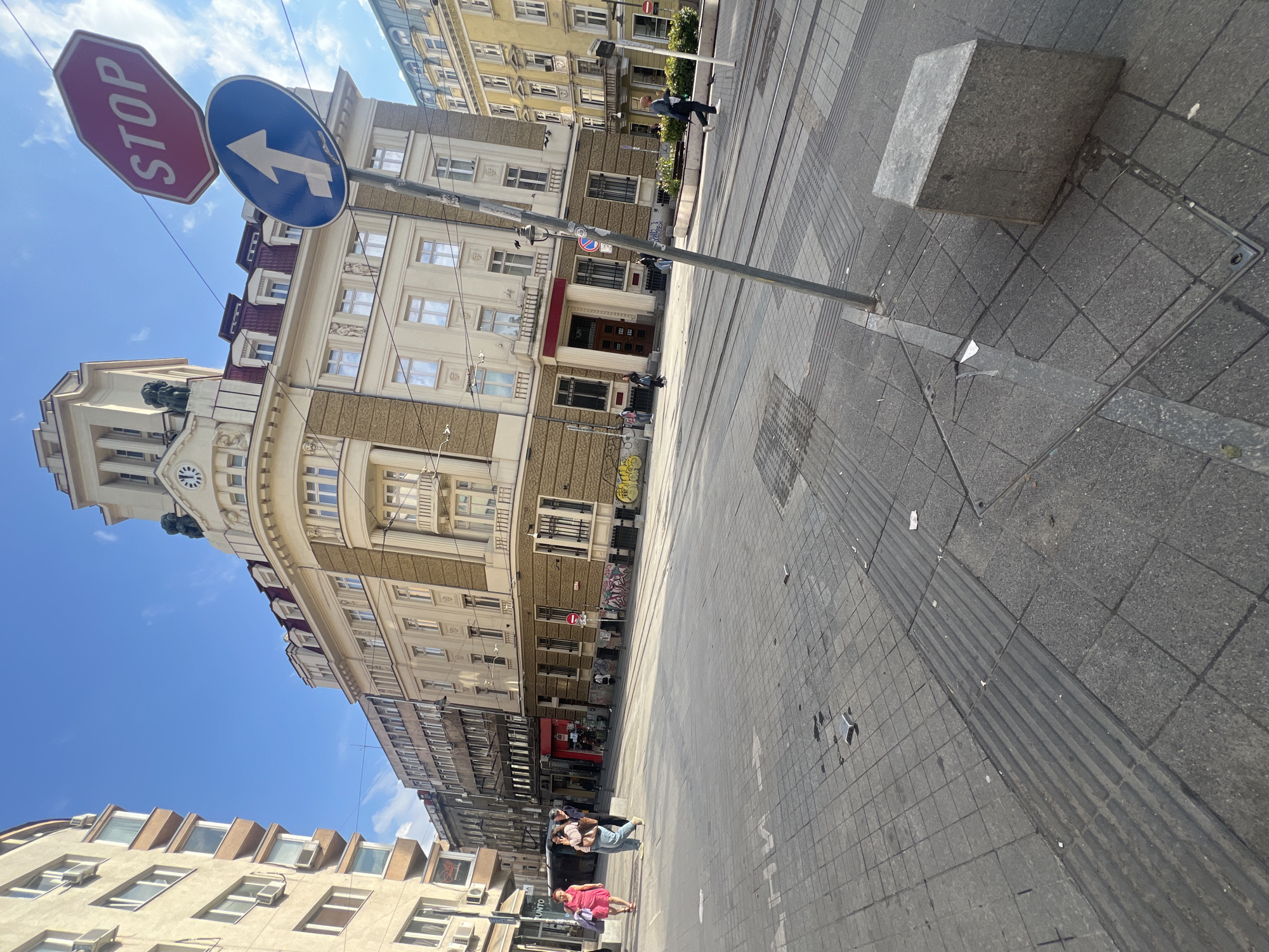 This bright, daytime photograph captures a lively street scene in a city, identified as Sofia, Bulgaria. The sun is high, casting clear shadows and illuminating the area under a vibrant blue sky with a few wispy clouds.
The street is lined with multi-story buildings on both sides. On the left, a prominent building showcases ornate architecture with classical details, including a large clock face, multiple windows, balconies, and a distinctive reddish-brown roof with dormer windows. Other buildings along the street feature various facades, some appearing residential and others commercial, with visible ground-floor shops and some instances of graffiti on lower walls.
In the mid-ground, a tall metal pole supports two distinct road signs. One is a red octagonal "STOP" sign with the word "STOP" in white capital letters. Below it, a blue circular sign features a white arrow pointing straight ahead, indicating a mandatory direction.
The street itself is paved with light and dark grey tiles, and tram tracks run down its length, suggesting a major urban thoroughfare. Sidewalks also feature tactile paving. Several people are visible: an individual in a light jacket walks on the right side of the street, while two people are seated on a low stone ledge or barrier on the left. Further down, another person in a pink dress stands on the sidewalk. A grey, rectangular stone block, possibly a seat or barrier, rests on the right sidewalk. The word "PUNTO" is faintly visible on a sign in the lower left. The overall scene depicts typical urban activity on a clear, sunny day.
