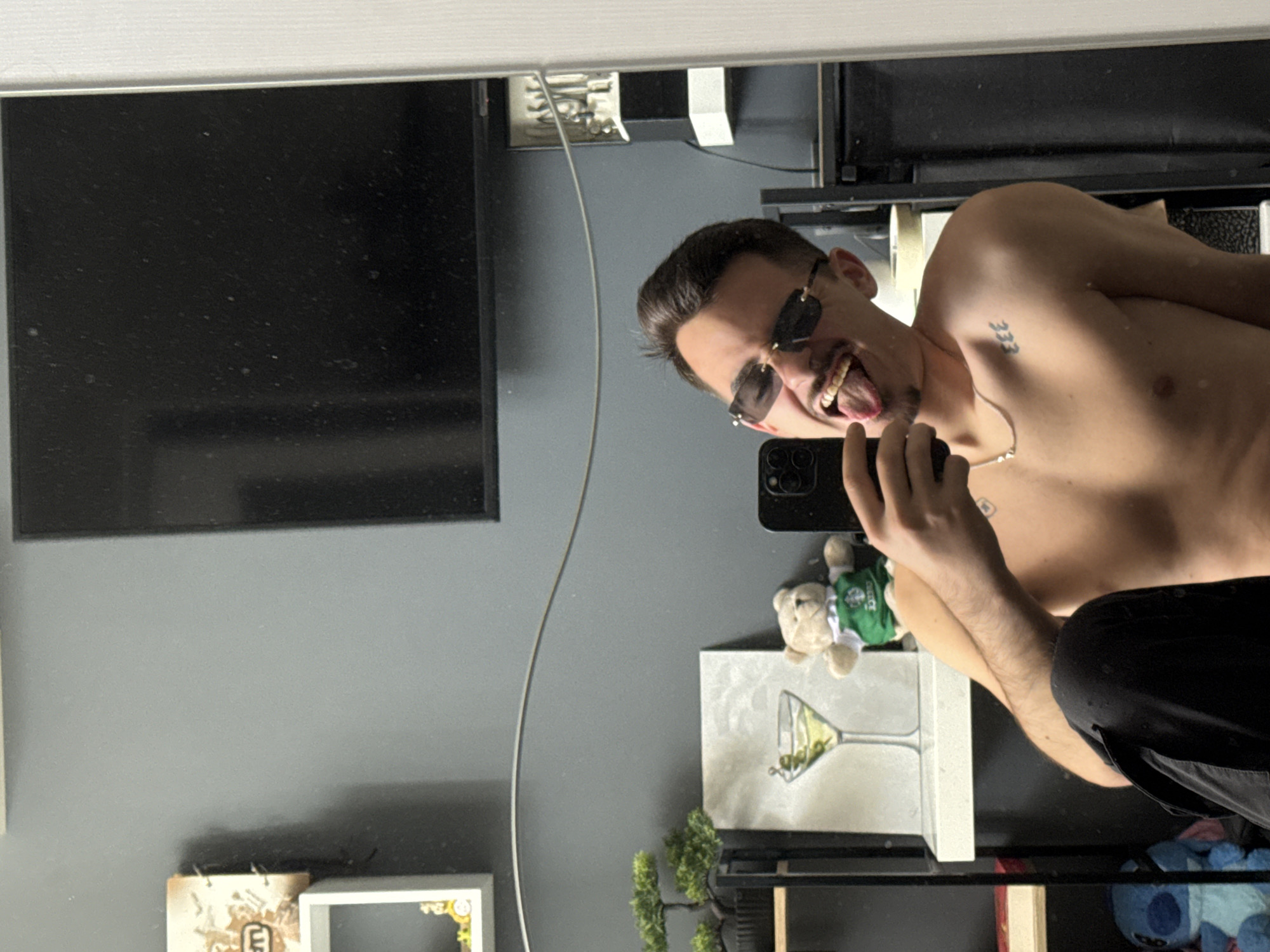 A young man is depicted taking a mirror selfie indoors. He is shirtless, capturing his head and torso, which occupy the right side of the frame. He sports dark, rectangular sunglasses and has dark hair. A tattoo of the number "333" is visible on his left shoulder. He holds a black smartphone in his right hand, using it to photograph his reflection. His expression is playful, characterized by a wide smile and his tongue sticking out.
The mirror's reflection reveals the room behind the man. A large, black television screen is prominent on the left side of the reflection. The walls are a neutral gray. Below the television, on what appears to be a shelf, several decorative items are arranged. These include a small teddy bear dressed in a green vest, a framed artwork featuring a martini glass with olives, and a small green plant resembling a bonsai. A thin white cable runs diagonally across the reflection. Further objects, such as a small book or calendar and a white empty frame, are also visible on the shelves. A light blue plush toy, possibly Stitch, is partially seen on a lower shelf.
The scene suggests an interior living space, likely a home in Sofia, Bulgaria, with a modern aesthetic. The man's demeanor is lighthearted and candid.