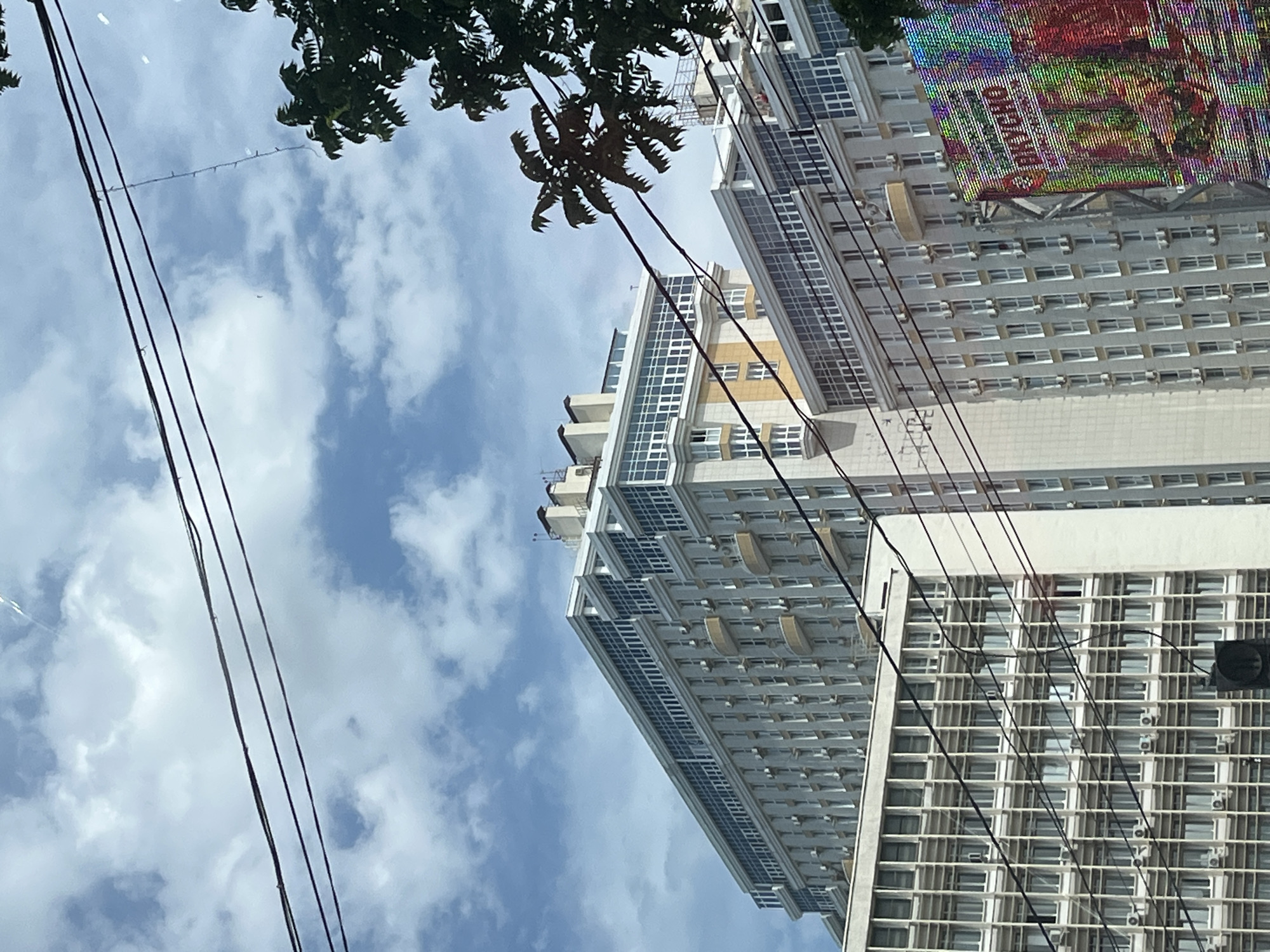 The image depicts an urban scene in Krasnodar, Russia, focusing on several multi-story buildings against a cloudy sky. Power lines are prominently strung across the foreground, obscuring parts of the buildings and sky. The architecture of the buildings varies, with some featuring modern glass facades and balconies, while others have a more utilitarian grid of windows. On the right side of the frame, a colorful, pixelated digital billboard is visible, displaying text that includes the Cyrillic word "ВКУСНО" (VKUSNO), meaning "delicious" or "tasty". A tree with dark green foliage encroaches on the upper right corner of the image. No individuals are clearly discernible in the scene.