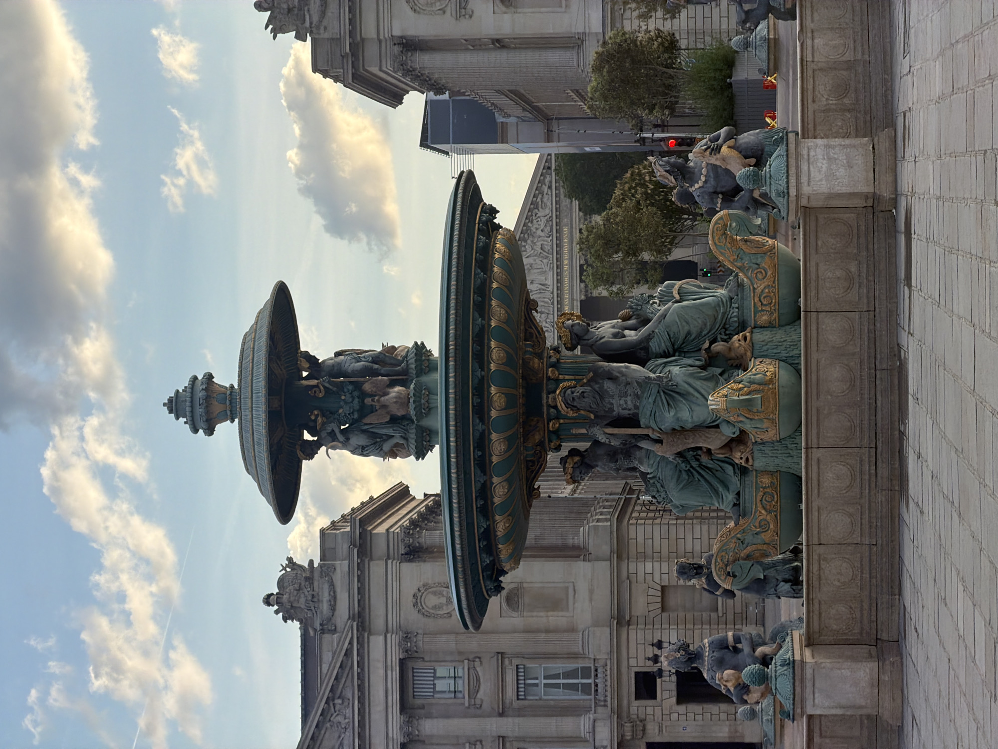 The image captures a view of the Fontaine Saint-Michel in Paris, France, set against a partially cloudy sky. The fountain is prominently featured in the foreground, showcasing its elaborate sculptural details and multi-tiered structure. The architecture of surrounding buildings, adorned with intricate designs, adds to the scene's historical ambiance. Paved surfaces are visible, hinting at the urban context and pedestrian traffic. Traffic lights and trees contribute to the urban landscape in the background. No people are clearly visible in the image. The focus is on the fountain and the surrounding architectural elements.