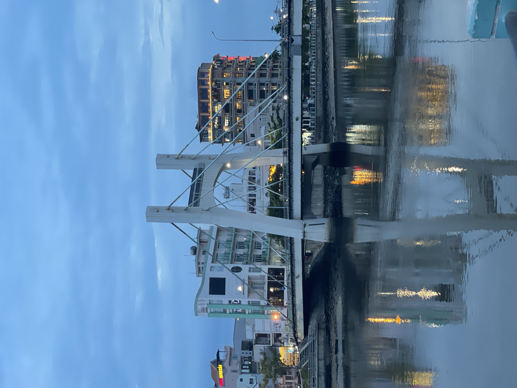 This media file captures an urban waterfront scene at dusk or early evening. The prominent subject is a modern, white pedestrian bridge that spans across a calm river or canal. The bridge features distinctive arch-like supports and a network of white suspension cables.
On both sides of the waterway, various multi-story buildings line the banks. Many of these buildings have illuminated windows, casting warm yellow and orange light, which contrasts with the deep blue twilight sky. On the left bank, a building is visible with a red sign clearly displaying the word "GOLDEN". Further along the same bank, another building has a dark facade with several lit windows. On the right bank, a building features a prominent red sign where the word "QUEEN" is partially legible. Streetlights are also visible, contributing to the urban illumination.
The calm surface of the water beautifully reflects the artificial lights from the buildings and the bridge, creating elongated, shimmering patterns that ripple across the blue-grey liquid. The sky above is clear and a rich blue, indicative of the transition from day to night. There is no discernible human activity taking place, lending a peaceful and tranquil atmosphere to the scene, emphasized by the interplay of artificial light and natural twilight.