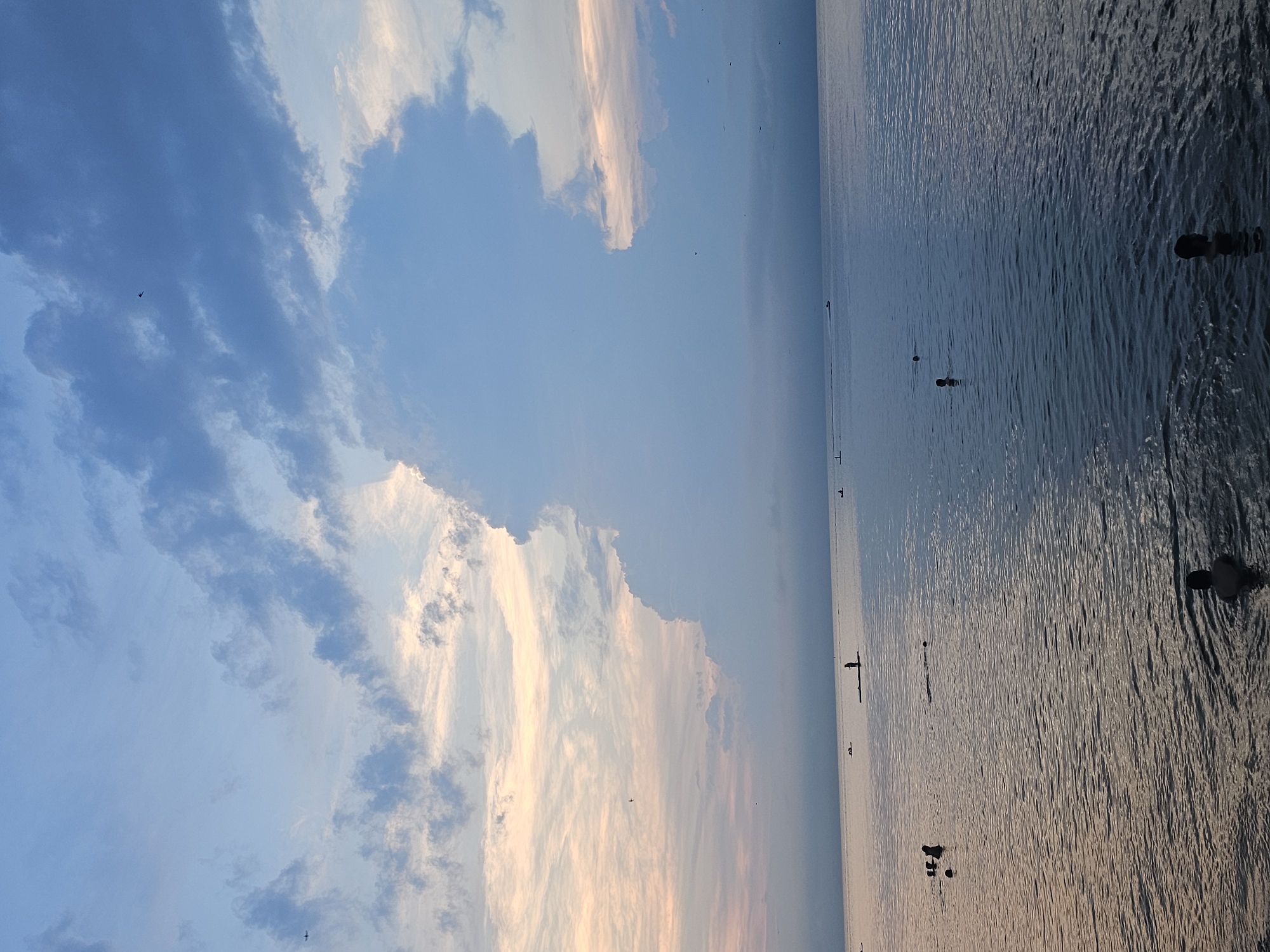 The image depicts a wide body of water, likely a sea or ocean, with a serene surface reflecting the sky. The sky is a canvas of blues and soft oranges, with dramatic clouds forming a large, towering mass on the right. Several small figures are visible in the water, some appearing to be swimming and others possibly stand-up paddleboarding. A distant boat can be seen on the horizon. The scene conveys a sense of tranquility and leisure, characteristic of a calm day at the beach or a waterfront location. Given the provided information, this scene could be located along the coast of Vladivostok, Russia, an area known for its scenic bays and proximity to the sea.
