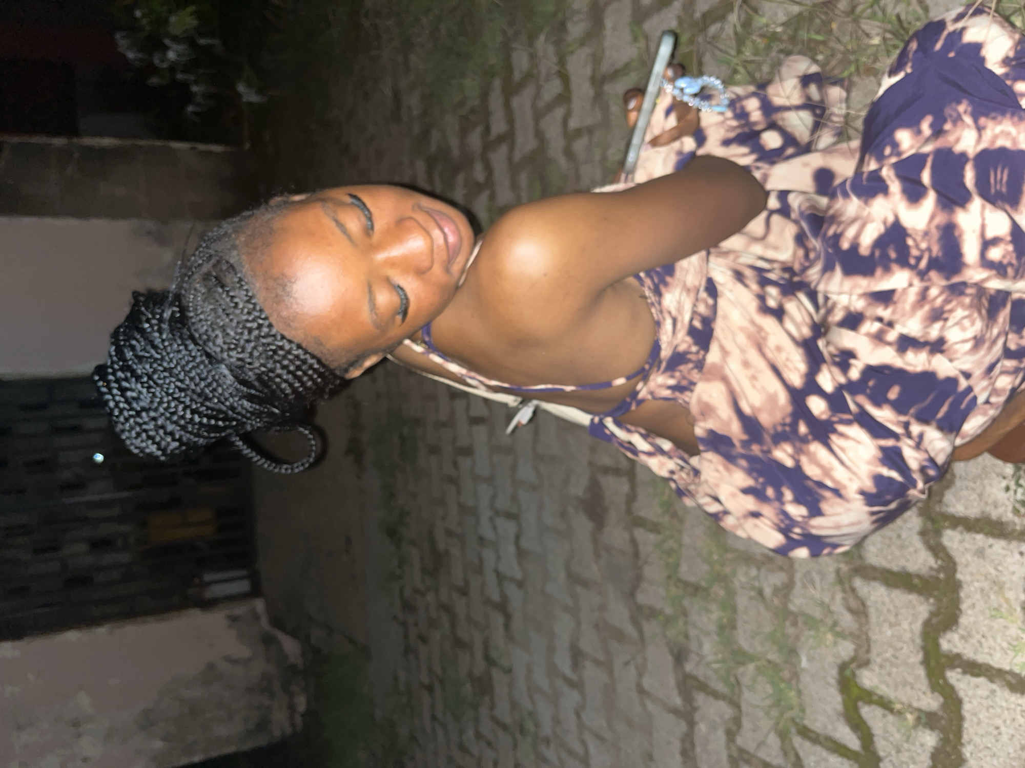 A young Black woman with her hair in a high braided bun is crouching on a paved outdoor area. She is wearing a tie-dye maxi dress with a pattern of dark purple and light brown/tan. Her head is tilted down and to her right, with her eyes closed and a slight smile on her face. She is holding a smartphone in her right hand, which is resting on her lap, and she is wearing a light blue bracelet on her left wrist. Behind her, a dark, textured wall and a metal gate are visible. The ground is paved with interlocking stones, and patches of grass are growing between them. The lighting suggests it is nighttime or dusk. The overall scene appears to be in an outdoor residential or public space, possibly a courtyard or walkway. While the specific location in Jos, Nigeria, is not visually identifiable, the scene depicts a common urban environment.