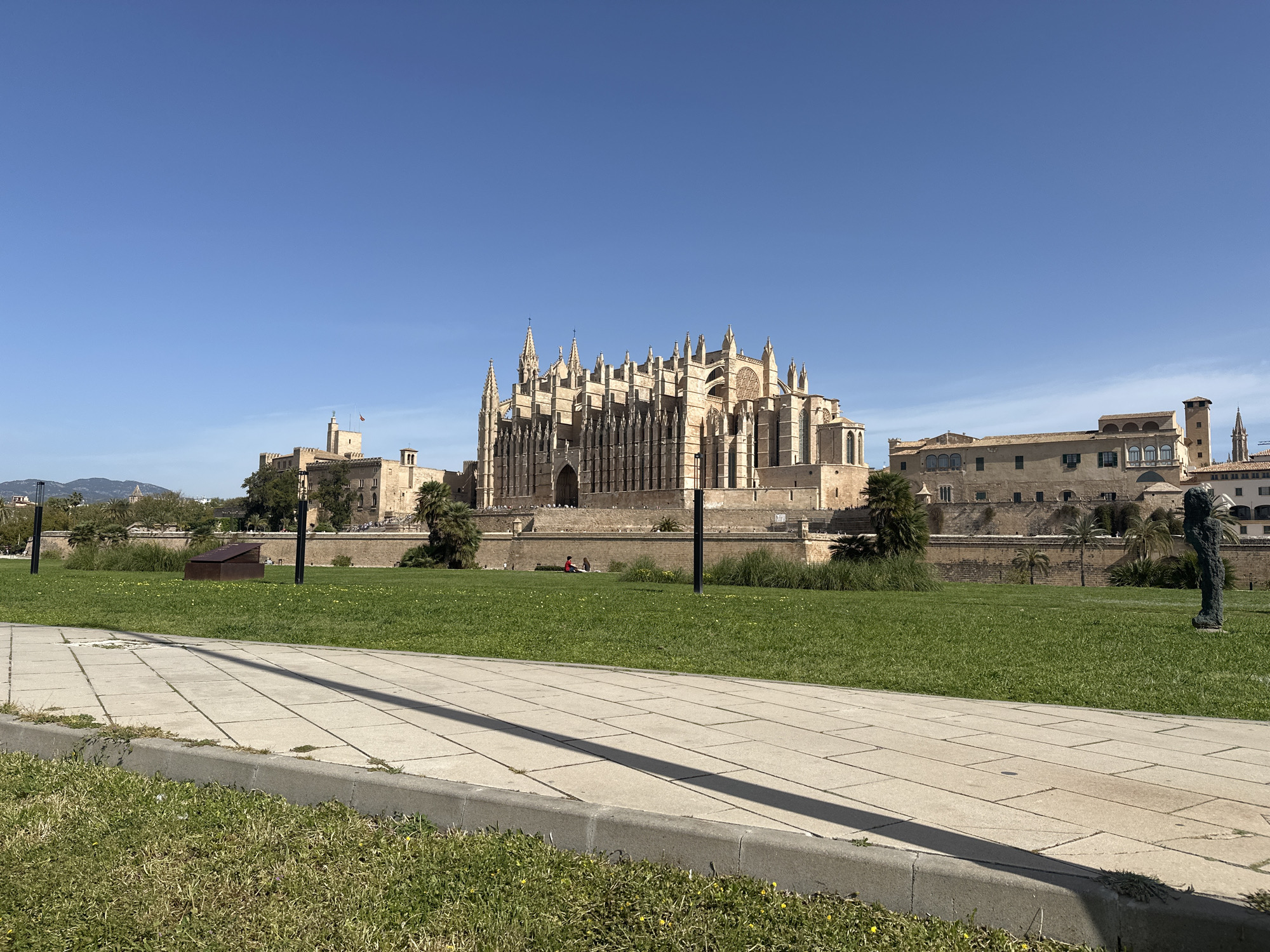 The image shows the Cathedral of Santa Maria of Palma, also known as La Seu, in Palma de Mallorca, Spain. The large Gothic cathedral dominates the background of the image. To the left of the cathedral is the Palau de l'Almudaina, a royal residence. A stone wall runs in front of both buildings. In the midground, a grassy area with trees and lamp posts stretches across the scene. A paved walkway is visible in the foreground. A statue stands in the right foreground. Two people are sitting on the wall near the cathedral in the distance. The sky is clear and blue.