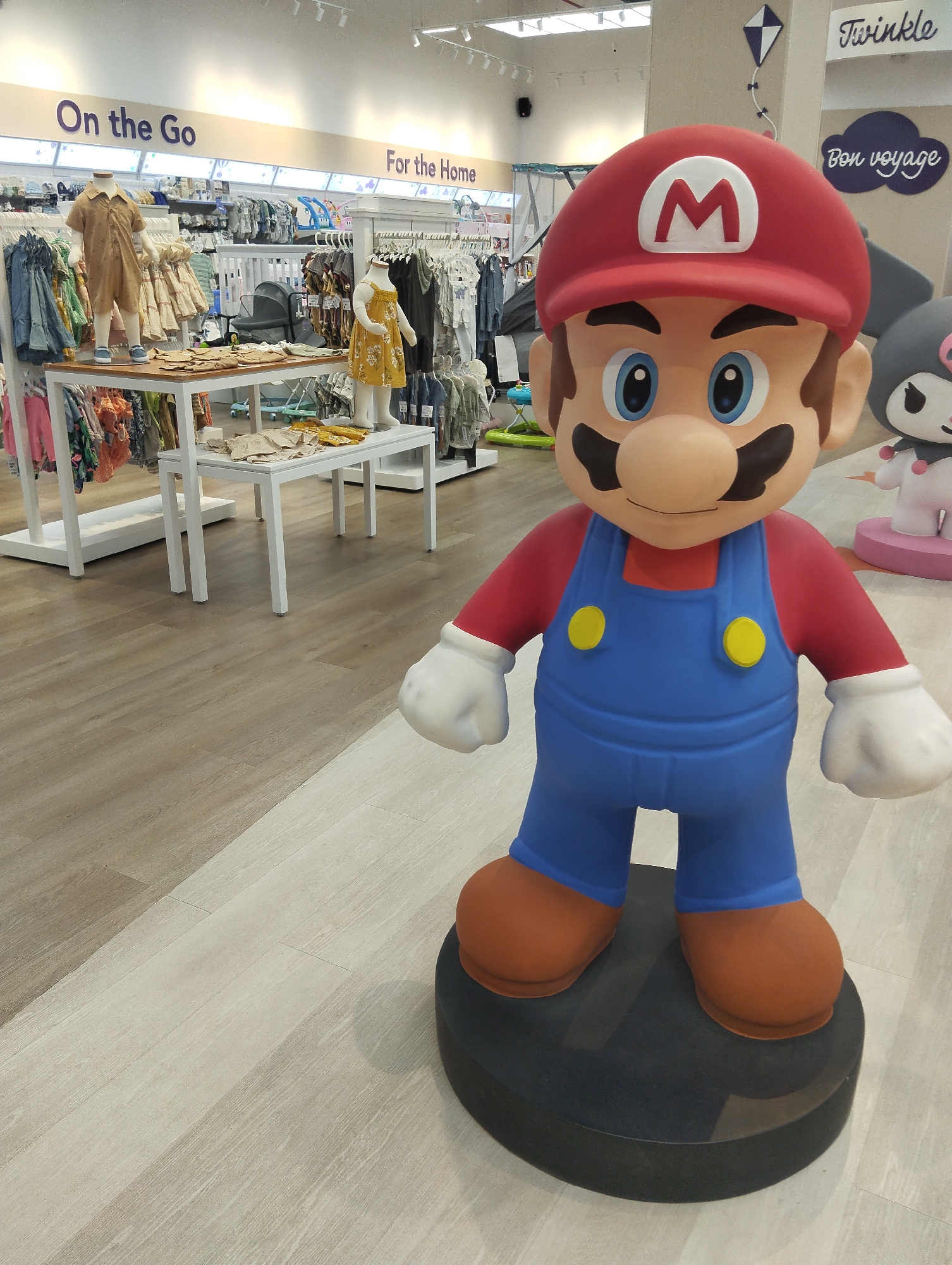 A large, colorful statue of the character Super Mario stands prominently in the foreground of an indoor retail store. Mario faces the viewer, depicted in his iconic red cap with a white 'M' emblem, red long-sleeved shirt, blue overalls with yellow buttons, white gloves, and brown shoes. He stands on a dark, circular base.
Behind Mario, the setting is a brightly lit children's clothing and accessories store. Racks are filled with various colorful garments, and two child-sized mannequins display a light brown romper and a yellow patterned dress. White display tables and shelves hold folded clothing and other items. In the background, further merchandise, including a baby stroller and other baby gear, is visible. Part of another cartoon character statue, resembling My Melody or Hello Kitty, is seen to Mario's right, resting on a pink platform.
The store features light wood laminate flooring that transitions to a paler shade near the Mario statue. Overhead, track lighting illuminates the space. Visible text on overhead signage includes "On the Go" and "For the Home" in purple lettering. Further text, also in purple, reads "Twinkle" and "Bon voyage," with the latter accompanied by a small kite graphic. No specific activity is taking place beyond the static display of merchandise and characters. The scene suggests a cheerful and organized retail environment, likely during daytime operating hours.