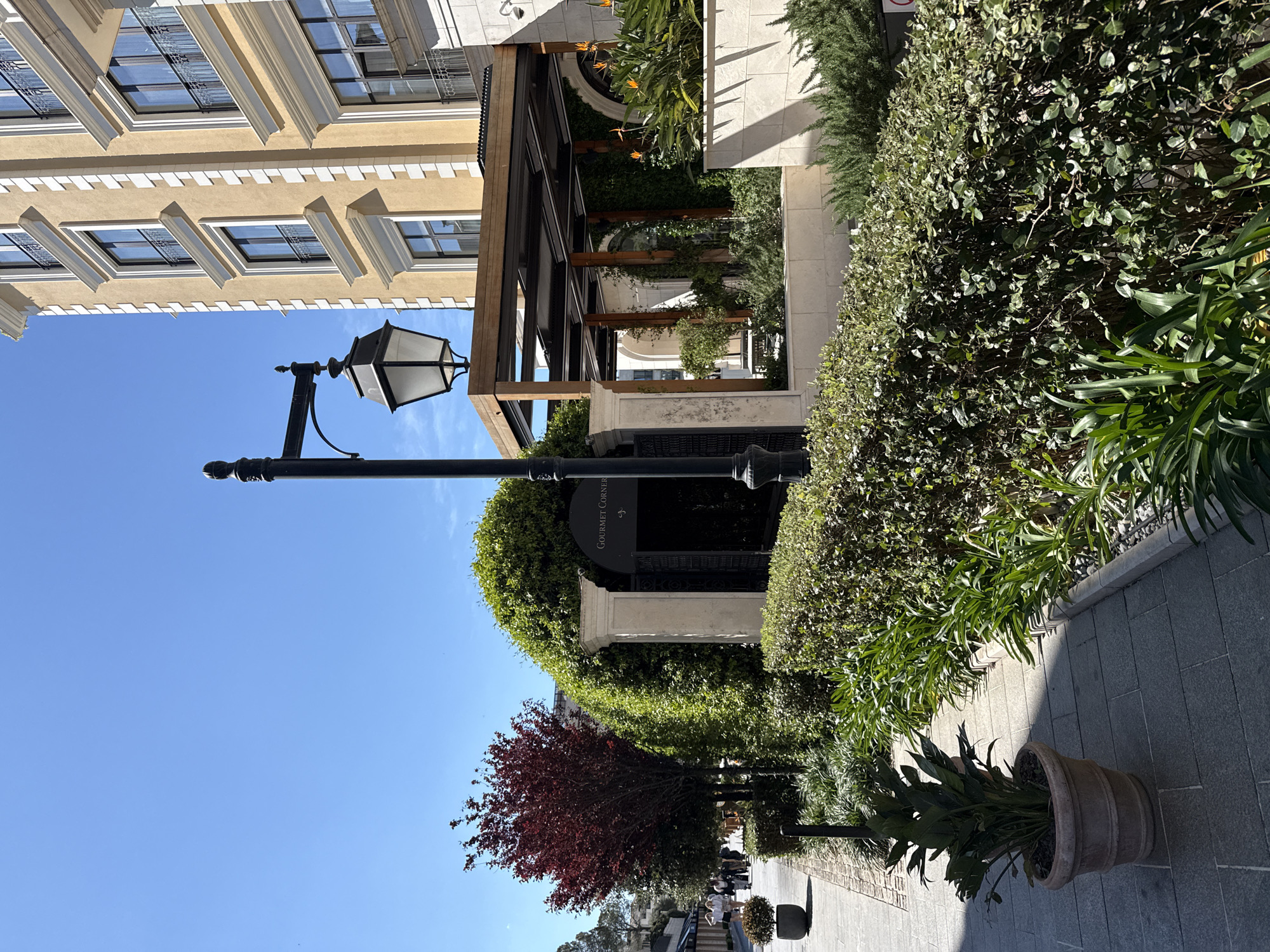 The image depicts an outdoor scene in Tivat, Montenegro. A sidewalk made of gray pavers is visible, leading towards an area with people walking. A large potted plant is present on the right side of the walkway. Landscaping includes a red-leafed tree and dense, manicured green hedges. A black lamppost stands tall next to the hedges. Above the hedge is an entrance, marked "GOURMET CORNER" on a sign. Beyond the entrance is a building with a wooden pergola and ornate stone architecture. The sky is a clear blue.