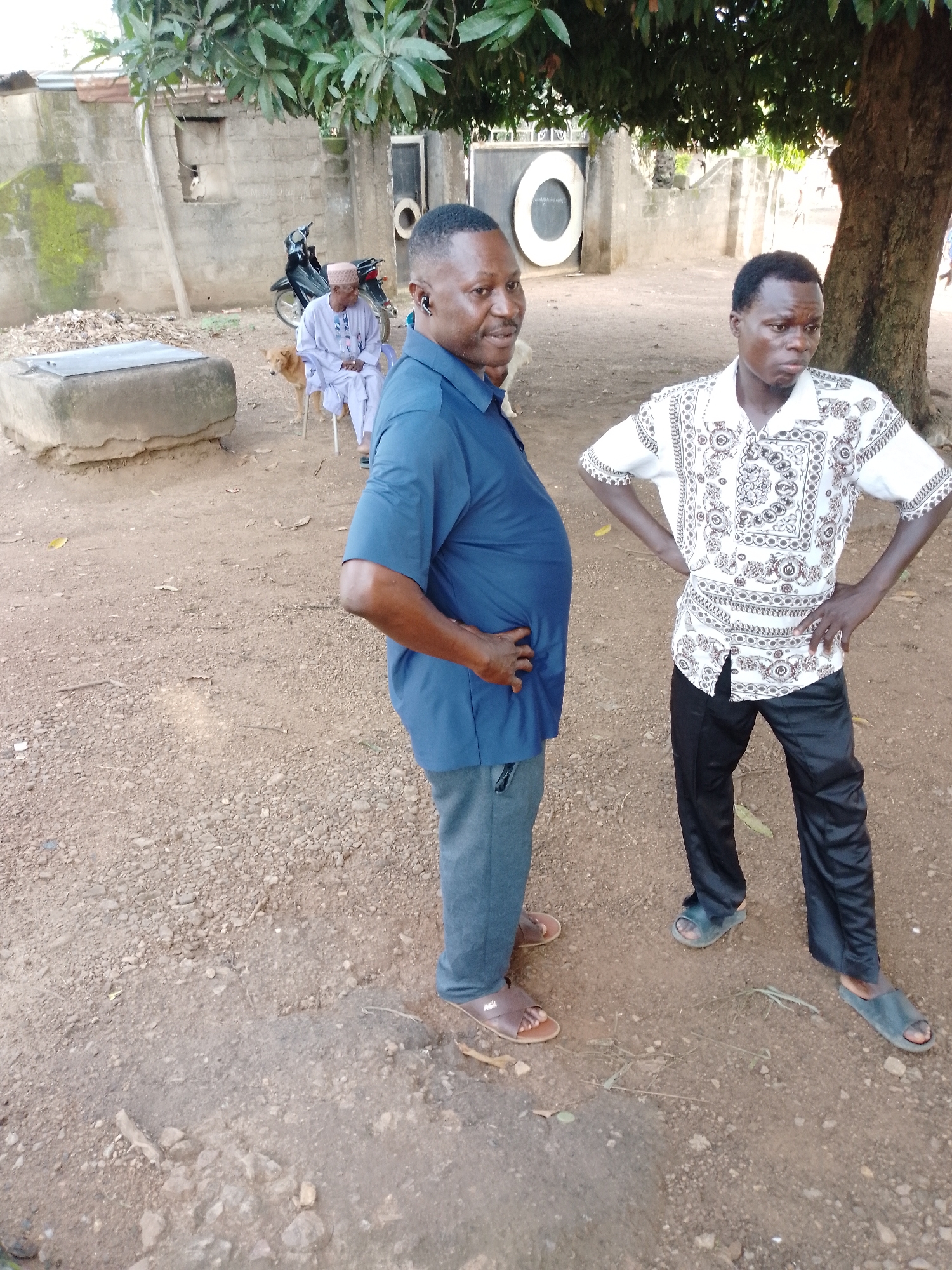 Two men are standing on a dirt ground, possibly outdoors in a rural or semi-urban area. The man on the left is wearing a blue short-sleeved shirt and grey pants, with his hands on his hips. He has a dark complexion and is wearing an earpiece in his left ear. The man on the right is wearing a white shirt with black intricate patterns and black pants, also with his hands on his hips. He has a darker complexion than the other man. In the background, a motorcycle is parked, and a man in a white traditional outfit is seated on a chair. There is also a dog near the seated man. A large tree is visible on the right side of the image, and in the background, there are stone walls and buildings. The ground is covered in loose stones and dirt.