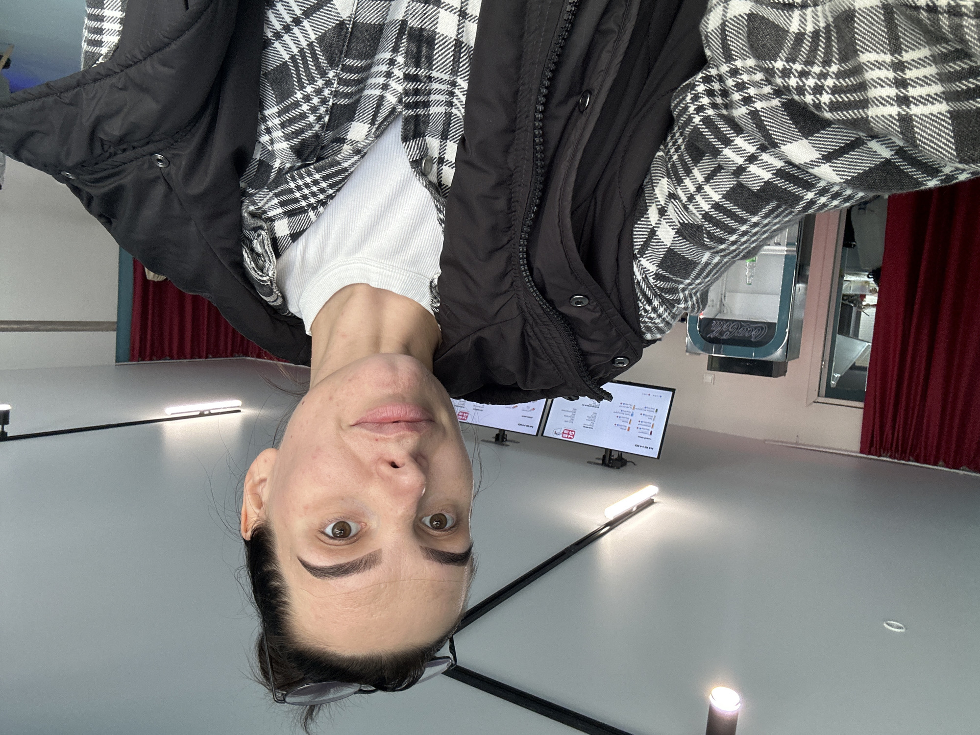 The image shows a medium shot of a woman with dark hair pulled up, wearing glasses perched on her head. She is looking directly at the camera, with her mouth slightly open. She is wearing a black puffer vest over a black and white plaid shirt, and a white t-shirt underneath. The background appears to be an indoor establishment, possibly a cafe or restaurant, with red curtains on the left side. In the background, there are digital menu boards displaying text. A vintage Coca-Cola vending machine is visible to the right of the woman. The overall lighting suggests an indoor environment. The location is identified as Chaikovsky, Russia.