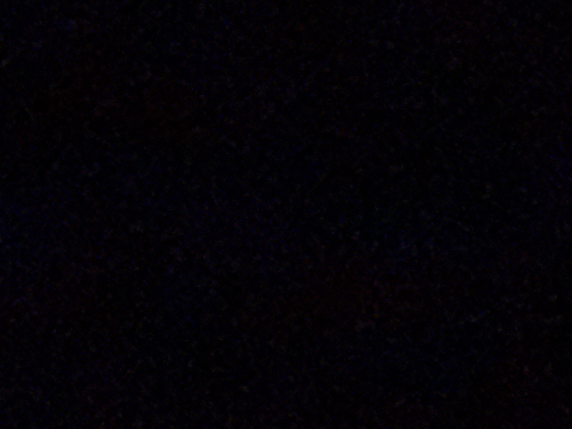 The image is a close-up, abstract view of a dark, textured surface, possibly fabric or a rough wall, with subtle hints of color, like deep red and blue, embedded within the blackness. There are no discernable objects, people, or environmental features that would allow for location identification within Navbahor, Uzbekistan, or any other city. The image appears to be a photographic study of texture and shadow.