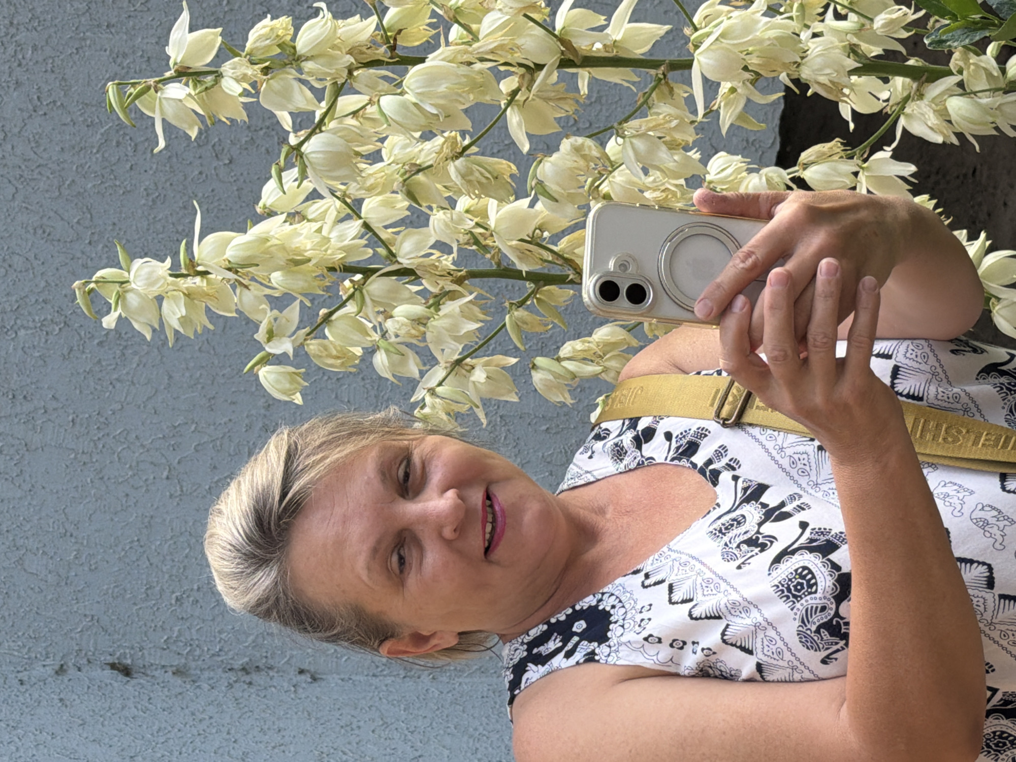 A middle-aged Caucasian woman with blonde hair styled up is standing outdoors in front of a blue-grey wall and a blooming yucca plant. She is holding up a silver smartphone in her right hand, appearing to take a selfie. She is wearing a sleeveless white top with a black and white elephant print and a yellow strap is visible over her right shoulder. Her mouth is slightly open as she smiles at the camera. The yucca plant has multiple stalks with clusters of white, bell-shaped flowers. The lighting suggests it is daytime, likely sunny. The scene appears to be in a backyard or garden setting.