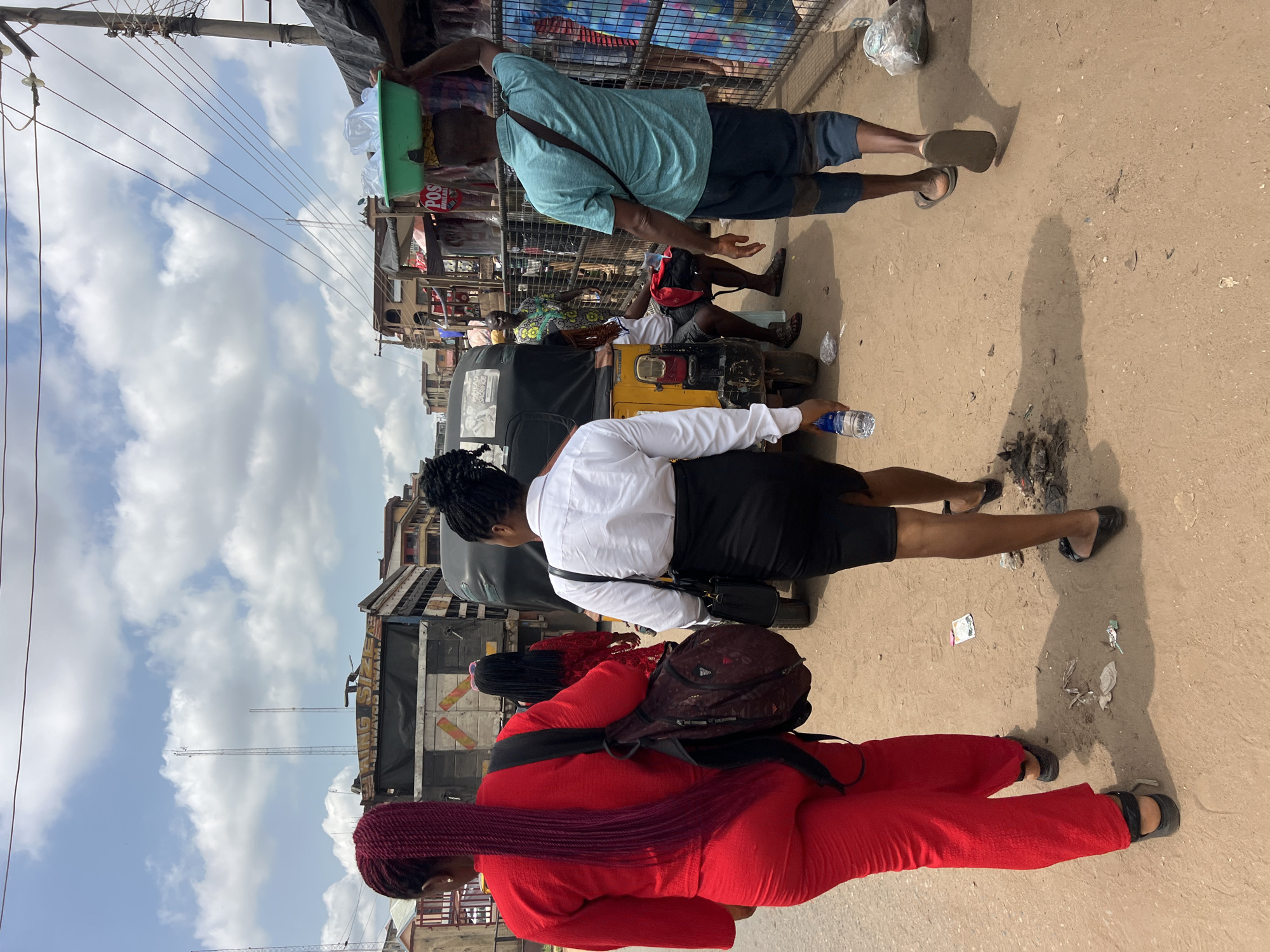 The image depicts a vibrant street scene in Nkpor, Nigeria, under a bright, cloudy sky, captured from a slightly elevated perspective. The setting appears to be a busy commercial area or market street, characterized by unpaved ground and various makeshift structures.
Several people are visible, indicating daily activity. In the foreground, a woman wearing a white long-sleeved shirt and a black skirt, carrying a black shoulder bag, walks to the left while holding a plastic water bottle. Behind her, another woman dressed in a flowing red outfit with long red braids also walks left, carrying an Adidas backpack.
Further into the scene, a yellow and black motorized tricycle, commonly known as a Keke Napep, is either parked or waiting. A person in a colorful patterned shirt and dark shorts is seated inside. To the right of the tricycle, a man in a light blue t-shirt and dark shorts carries a green plastic basin on his head, walking towards the right side of the frame. Another man's legs in dark shorts and sandals are visible on the dusty ground.
The ground is an unpaved, sandy-brown surface scattered with debris, including plastic items and what looks like discarded refuse or burnt material. Strong, well-defined shadows are cast on the ground, indicating bright daylight, likely mid-morning or early afternoon, despite the presence of white cumulus clouds in the sky. Overhead, a network of utility lines stretches across the frame, connected to visible poles. In the background, various market stalls and commercial buildings with simple constructions and corrugated iron roofs line the street. A red sign with white lettering displaying "POS" is clearly visible, with additional text "S_BLE" partially legible below it, suggesting a point-of-sale service. The scene captures the essence of everyday life and commerce in a Nigerian urban environment.