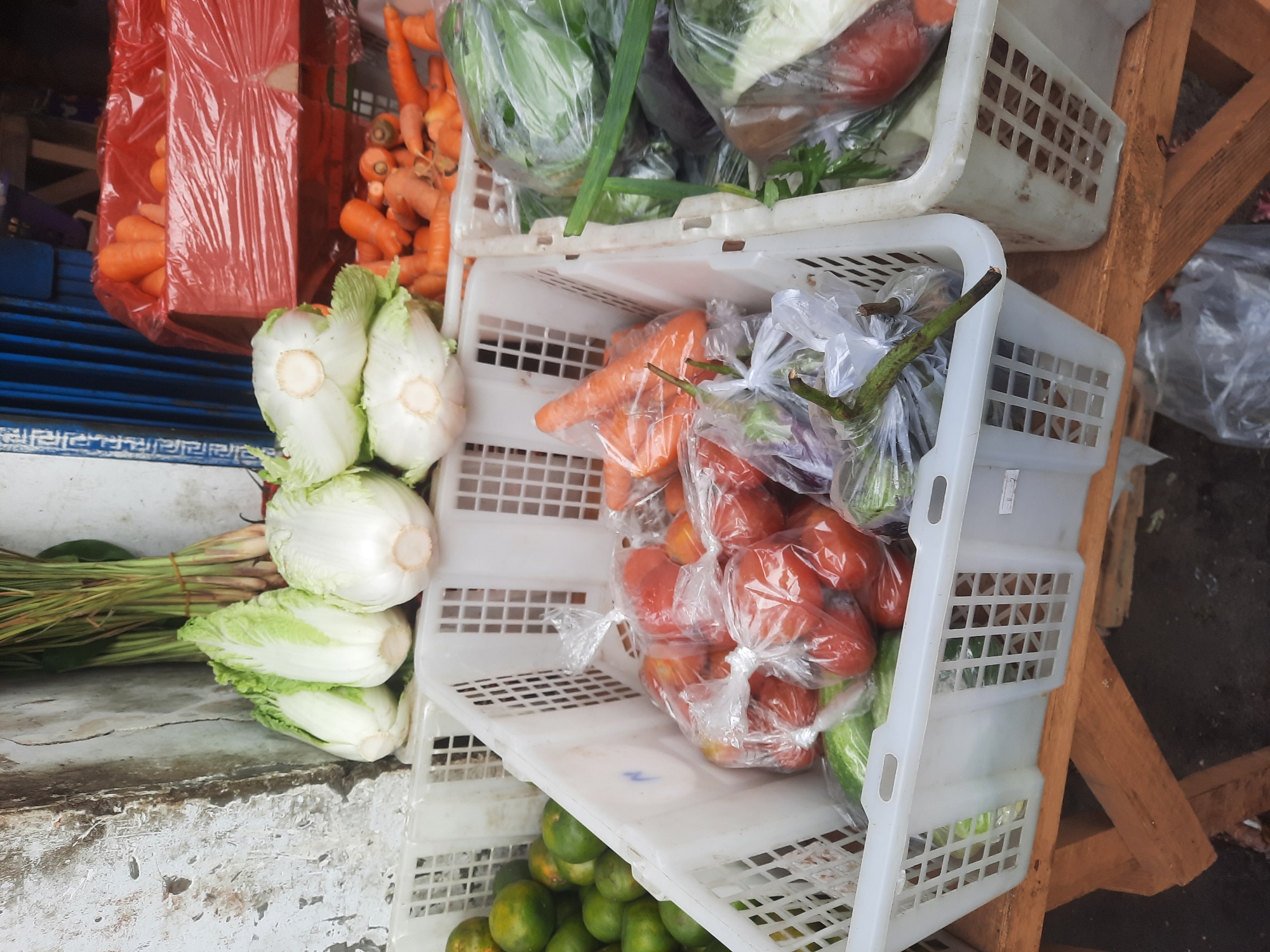The image depicts an outdoor market stall in North Jakarta, Indonesia, likely on a street or sidewalk. In the foreground, several white plastic crates are filled with fresh produce. The largest crate in the center contains a large quantity of tomatoes, most of which are bagged in clear plastic. Also visible in this crate are several green cucumbers. To the left, another crate holds a pile of small, green citrus fruits, possibly limes or small oranges. A third crate, partially visible on the right, contains green leafy vegetables, likely cabbage or lettuce, and some green onions. Behind these crates, other vegetables are visible. In a red bin covered with red plastic sheeting, a large pile of carrots is visible. To the left of the carrots and behind the white crates, several heads of Napa cabbage are arranged. A bundle of lemongrass stalks is also visible in the background. The stall appears to be set up on a wooden structure, possibly a market table or stand. The background shows a weathered, textured wall on the left and a blue shuttered structure on the right, suggesting a storefront or building facade. The overall scene is typical of a vibrant local market in Indonesia.