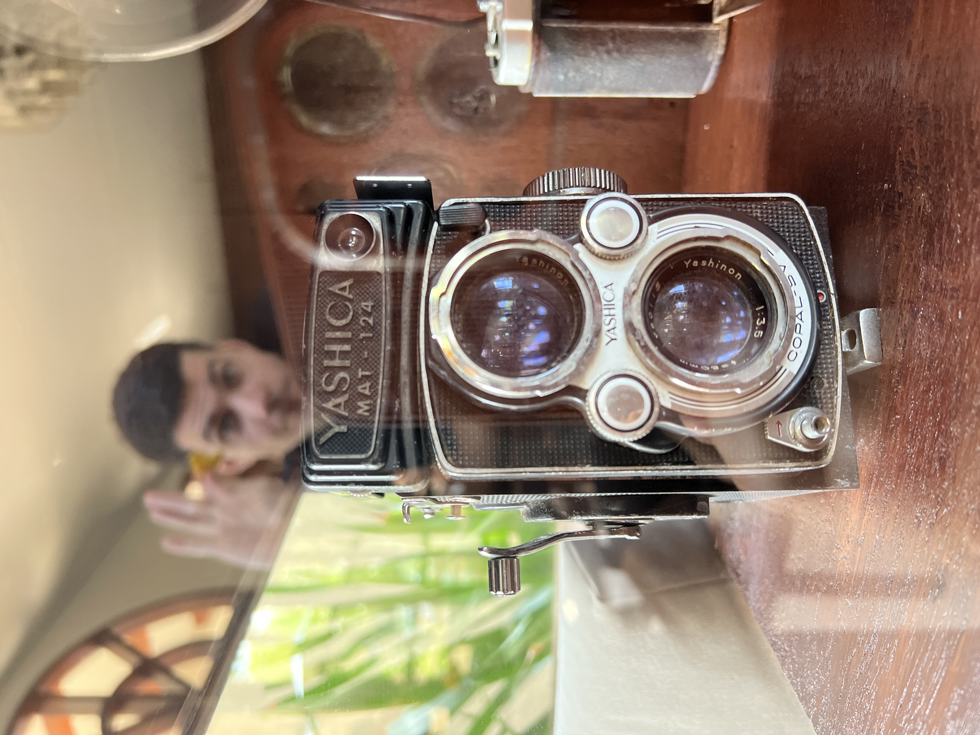 A medium close-up shot captures a vintage Yashica Mat 124 twin-lens reflex camera, positioned centrally on a polished wooden surface. The camera is the primary subject, with its lenses and branding clearly visible. In the background, a man is visible in a reflective surface, appearing to be taking a selfie with a yellow object in his hand. The man is out of focus and his facial features are not distinct. The environment appears to be indoors, with some foliage visible through a window or glass enclosure to the left. The overall lighting is dim, with the camera being well-lit. The context of Ubud District, Indonesia, is not directly inferable from the visual elements of the image itself.