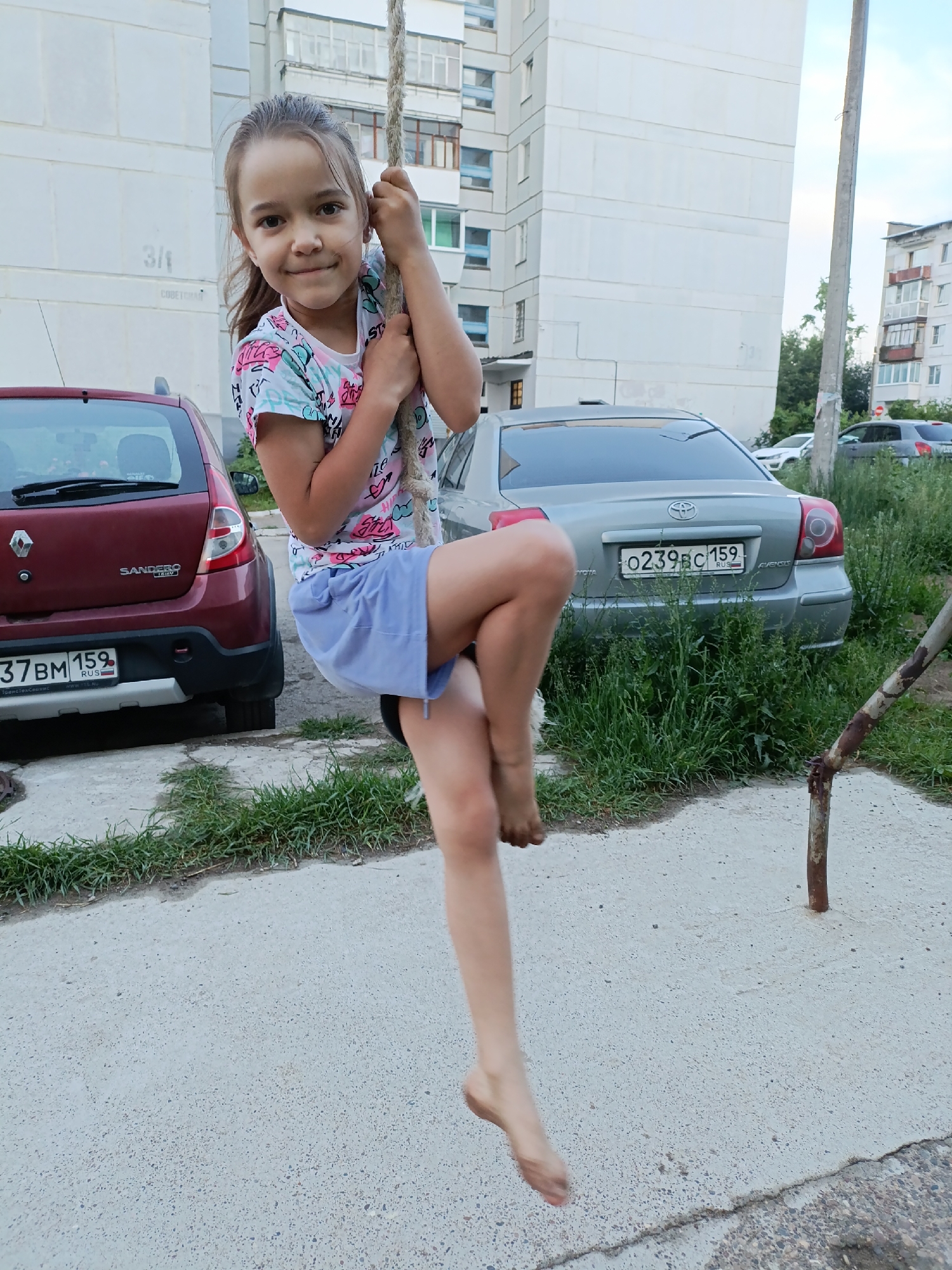 A young girl with dark hair pulled back is captured mid-air, swinging on a thick rope. She is wearing a white t-shirt with a colorful pattern of pink, blue, and black designs, and light blue shorts. Her bare feet are visible as she hangs from the rope, one leg bent and the other extended. She has a slight smile and is looking towards the camera.
The setting appears to be an outdoor urban environment, likely a residential courtyard. Behind her, a light-colored apartment building with multiple windows stands tall. To the left, the rear of a red Renault Sandero car is visible, with a license plate reading "37BM 159 RUS". To the right, a silver Toyota Avensis is parked, its license plate reading "0239BC 159 RUS". Tall green grass and some weeds grow between the cars and the concrete ground. A rusty metal pole is also present in the foreground on the right side. The lighting suggests it is daytime, possibly late afternoon or early evening, with soft light conditions. The overall mood seems playful and carefree.