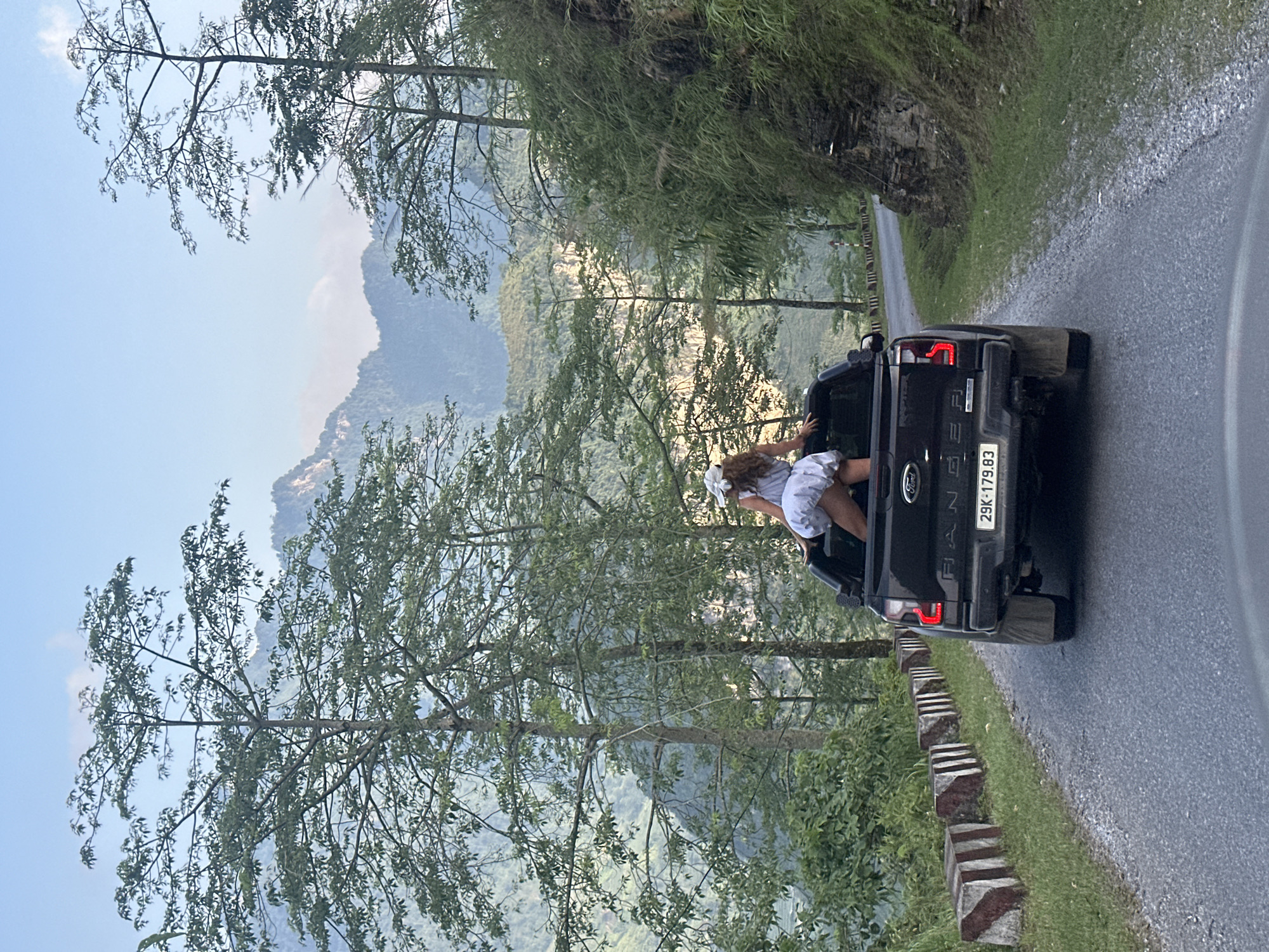 A black Ford Ranger pickup truck is parked on a curving paved road in Đường Thượng Commune, Vietnam, surrounded by a mountainous, forested landscape. The license plate number is 29K-179.83. A person wearing light-colored clothing is visible inside the truck bed. Trees and other vegetation frame the scene.