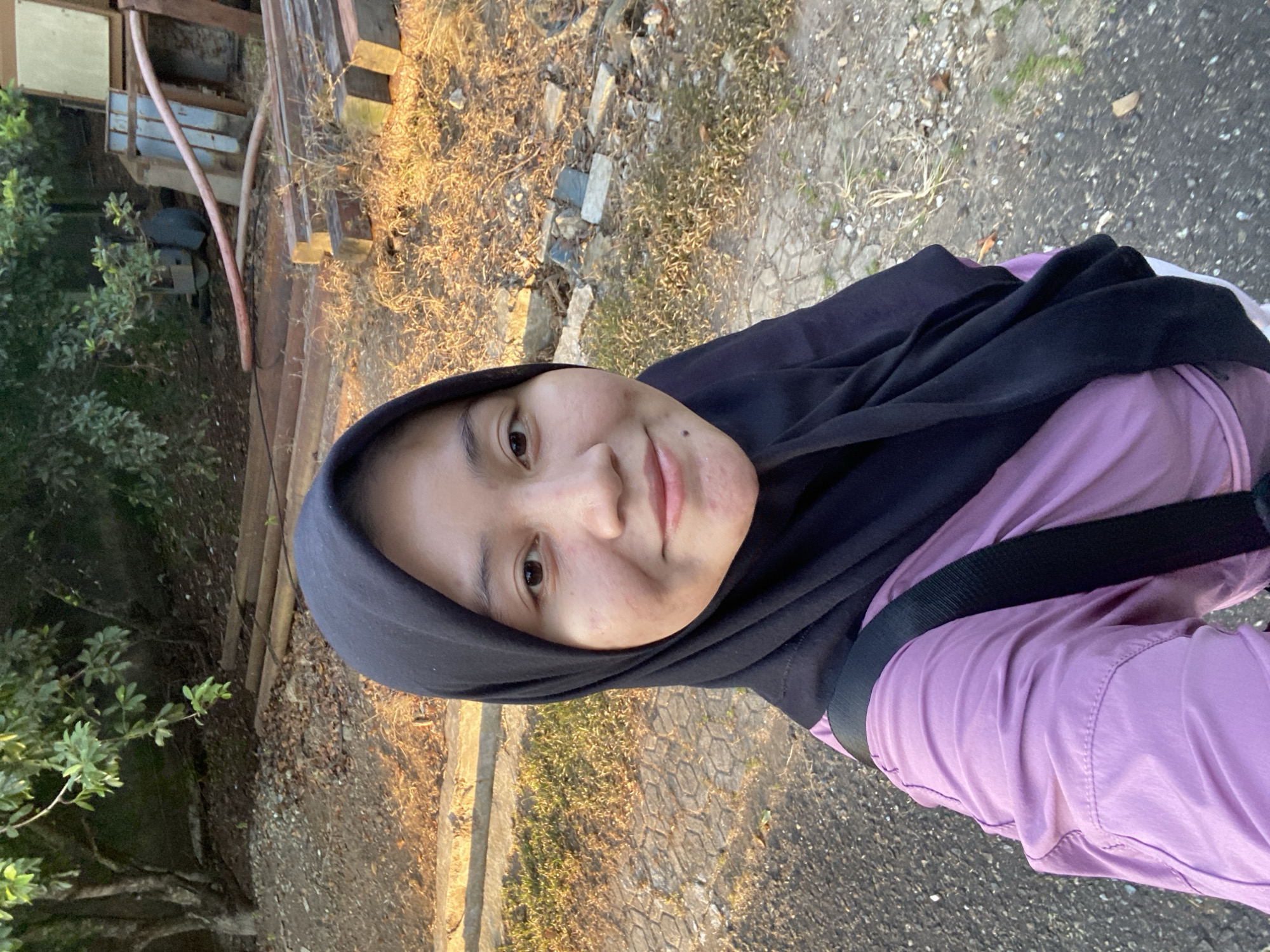 A young Indonesian woman wearing a black hijab and a lavender shirt is taking a selfie outdoors. She is smiling and looking directly at the camera. The background suggests an outdoor area with some trees, dry grass, and what appear to be construction materials, possibly lumber and pipes, scattered on the ground. The lighting indicates it is either early morning or late afternoon, with warm sunlight casting shadows. The terrain is a mix of asphalt and textured paving. The scene does not provide enough information to definitively place it within the city of Banjarbaru, Indonesia, beyond the general impression of an outdoor, somewhat undeveloped area common in many parts of the country.