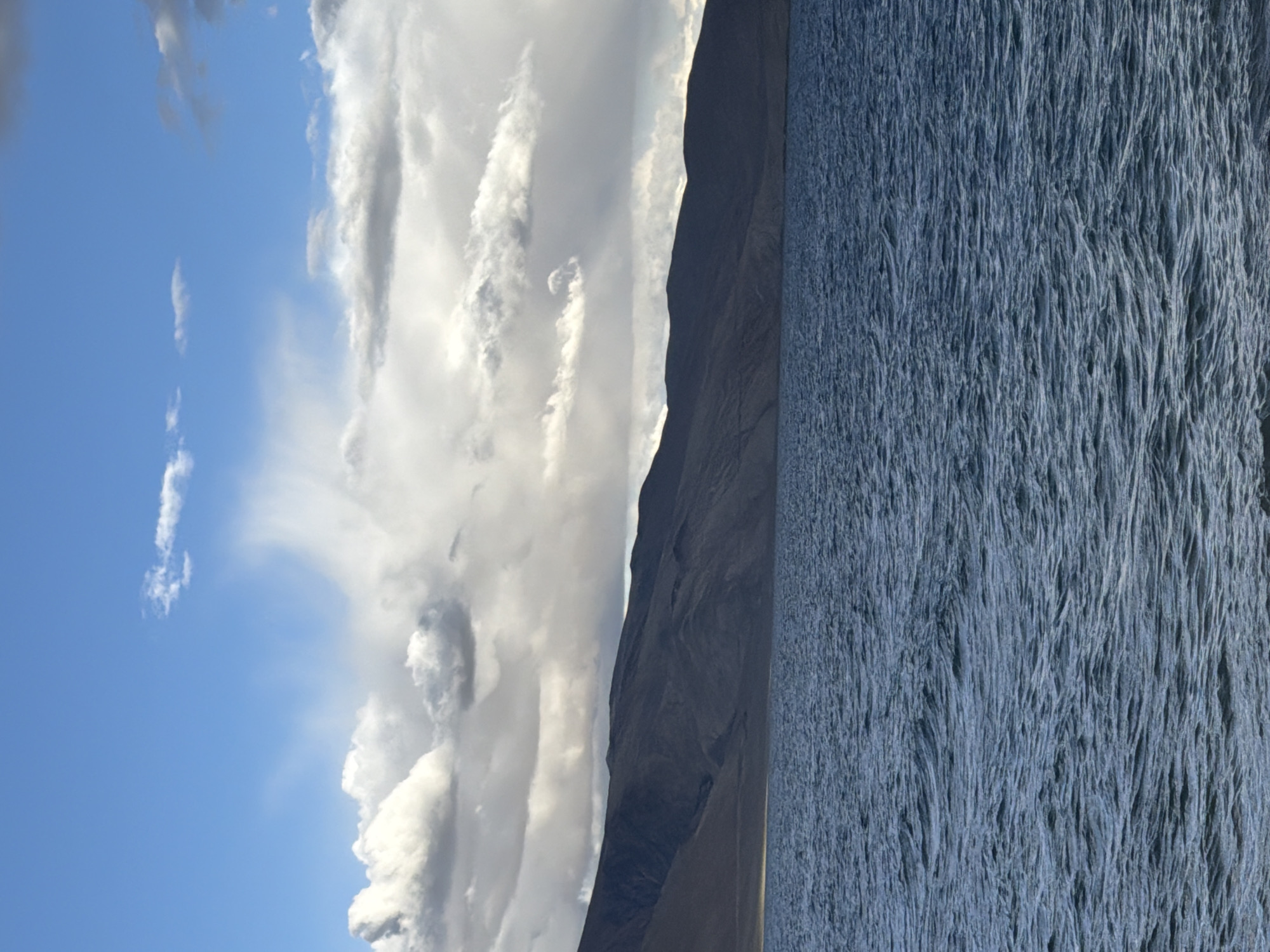 An image captures a body of water with waves under a cloudy sky. In the background, dark, undulating mountains rise from the shoreline. The scene appears to be a natural landscape, possibly a lake or a large river, given the presence of mountains and the expanse of water. The sky is a mixture of blue and white clouds, with some areas appearing brighter than others, suggesting varying light conditions. There are no people or man-made structures visible in the frame. The overall impression is one of a serene, natural environment.