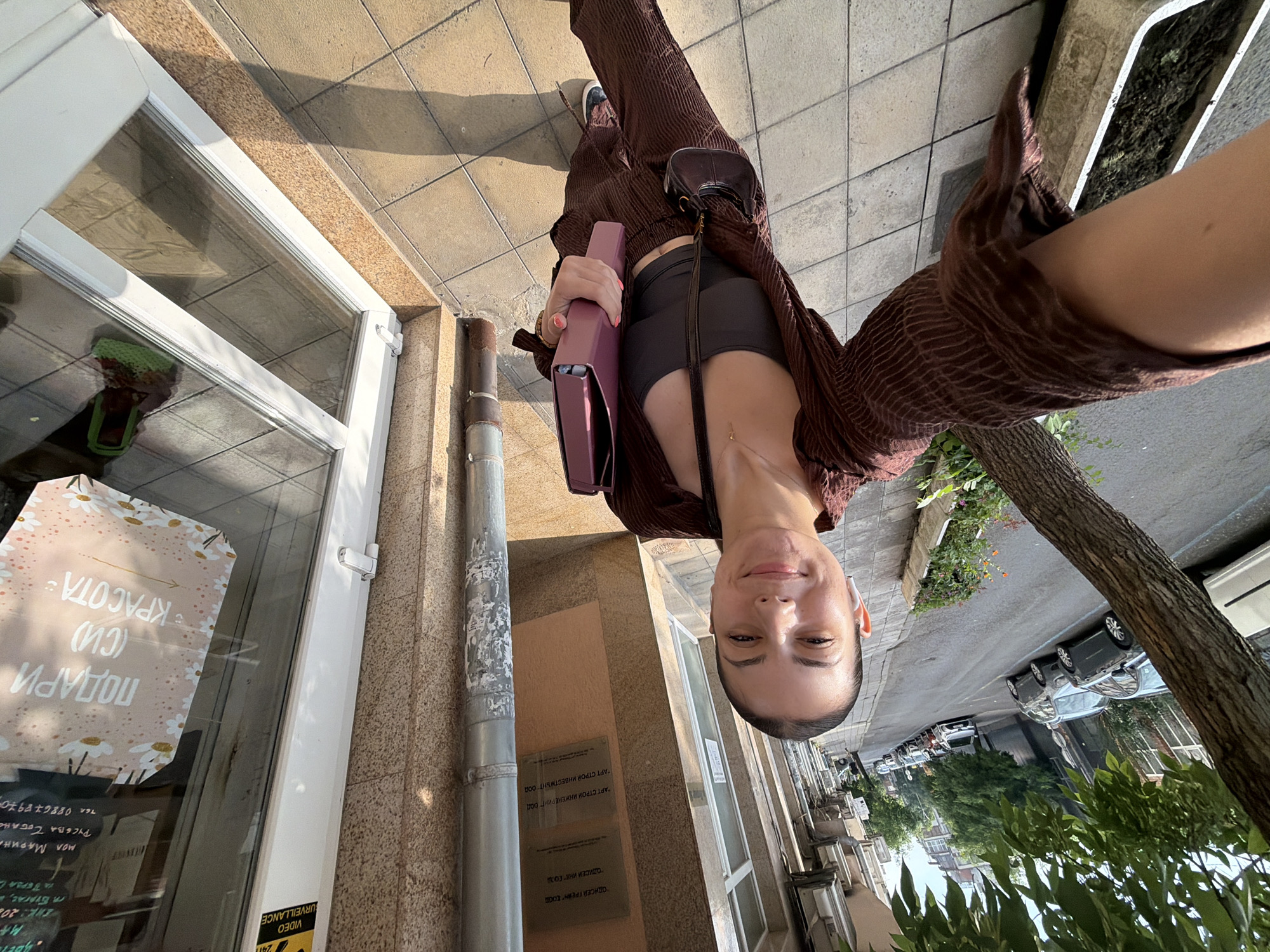 The image is a low-angle, upside-down selfie of a young woman smiling directly at the camera. She is holding a purple folder or clutch bag in her left hand and is dressed in a dark crop top, dark shorts, and a brown, crinkled long-sleeved shirt worn open. A small, dark shoulder bag and a delicate necklace are also visible.
The scene depicts an urban street in Burgas, Bulgaria, under bright, clear daylight, evidenced by distinct shadows. The foreground features a tiled sidewalk and the facade of a building with large glass windows. Reflected in the windows is a promotional sign, which reads, "ПОДАРИ (СИ) КРАСОТА" (Gift (yourself) beauty), along with "МОЛ МАРИНА", "РУСЕВА ТОБАНО", a phone number "088678970", and "г. Бургас". A yellow sign with "VIDEO SURVEILLANCE 24H" is also visible near the window frame.
On the building's wall, a plaque displays company names in Cyrillic: "ОДИСЕЙ ГРЕЙНТ OOD", "ОДИСЕЙ ИНА ЕООД", "АРТ СТРОЙ ИНЖЕНЕРИНГ ООД", and "АРТ СТРОЙ ИНВЕСТМЪНТ ООД". A metal drainpipe runs vertically alongside the building. In the background, a street is visible with several parked cars, flanked by additional buildings and green foliage from trees and bushes. The overall atmosphere is casual and sunny.
