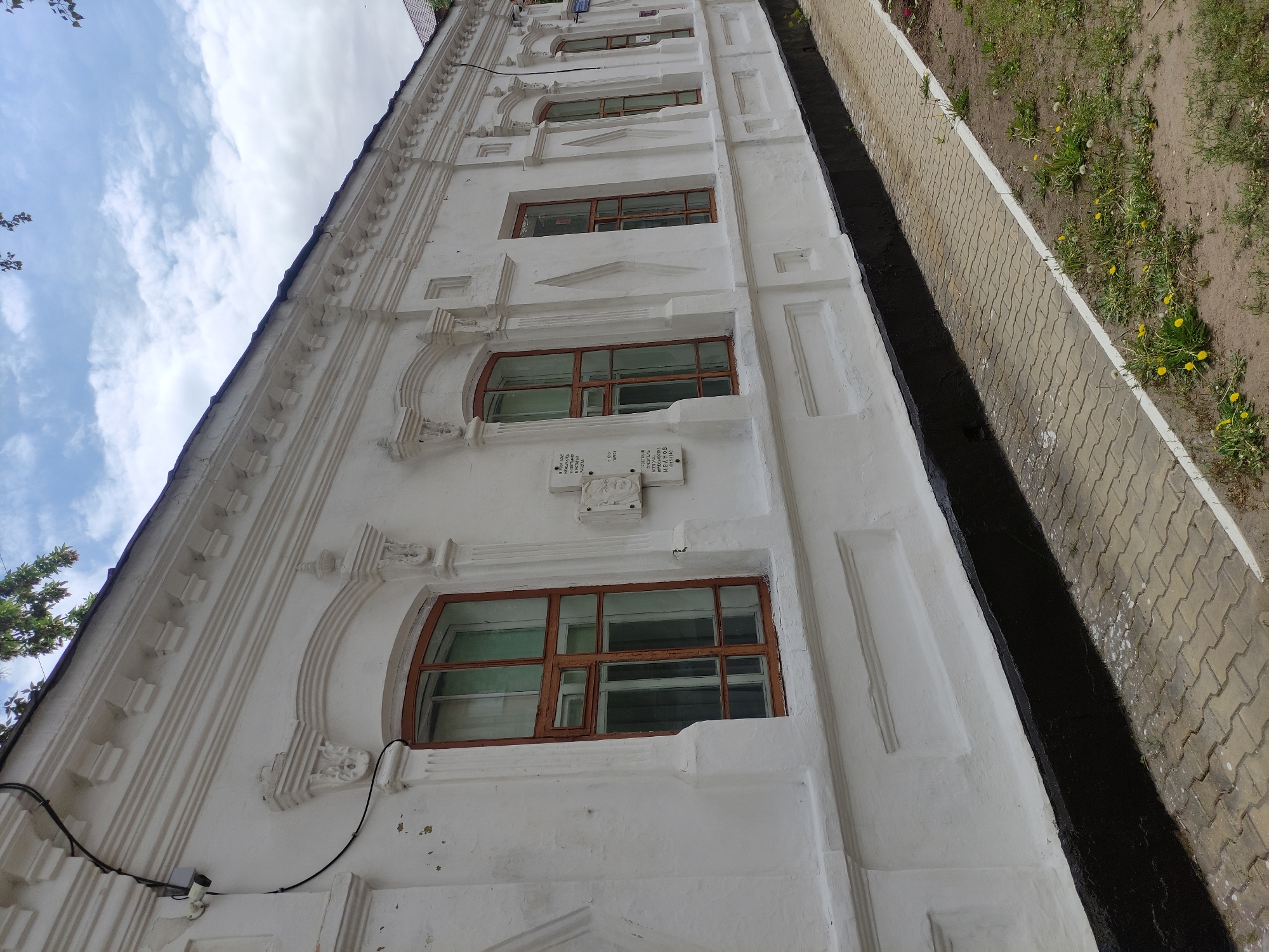 The image shows a low-angle view of a white building with several arched windows with brown frames. There are decorative architectural details along the building's facade. A black gutter runs along the building's right side, adjacent to a sidewalk paved with light-colored bricks. Beyond the sidewalk, there is a patch of dirt with vegetation, including some yellow flowers. Above, there is a view of a cloudy sky. Based on the information embedded in the image metadata and visible on plaques, the building is located in the city of Pavlodar, Kazakhstan.