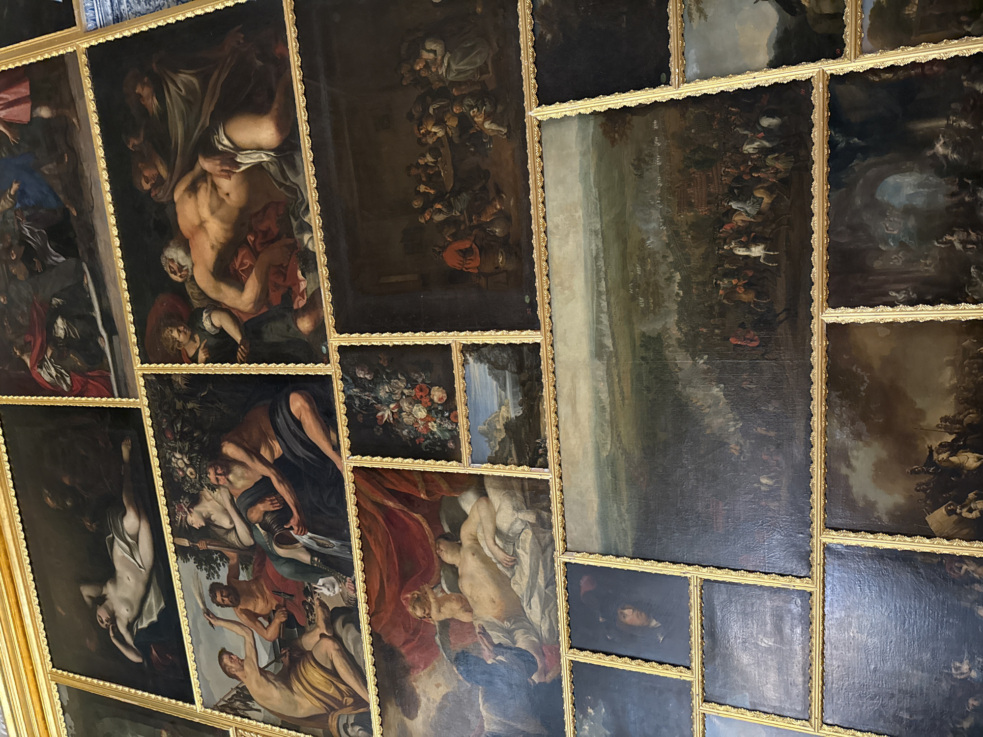 The image depicts a wall adorned with a collection of oil paintings, likely within a grand interior space. The paintings are framed in ornate gold moldings and arranged in a grid-like fashion, with varying sizes and orientations. The subjects of the paintings are diverse, featuring:
In the upper section, there are several paintings with human figures. One shows a reclining nude female figure draped in white fabric, possibly a mythological or allegorical scene. Another features a group of men and women gathered around a table in an interior setting, appearing to be engaged in conversation or a meal. There is also a painting of a sleeping muscular male figure being attended to by others, possibly a biblical or mythological depiction of a hero or god. Another painting shows a group of figures with one in the foreground holding a staff and another pouring liquid from a vessel.
The middle section of the wall showcases more paintings. A large horizontal painting in the center depicts a historical battle scene, with numerous soldiers on horseback and foot engaged in combat across a landscape. To the left of this battle scene, there is a portrait of a person wearing a hat and a dark coat, with a somber expression. Above the battle scene, there are smaller paintings, including one with a still life of flowers and fruit, and another depicting a landscape with a castle or fortress by a body of water.
The lower section of the wall continues with more paintings. Some appear to be portraits, while others show scenes with figures and architectural elements, possibly depicting mythological or religious narratives. One painting at the bottom right seems to portray a scene with figures in the foreground on horseback, with a dramatic sky above.
The overall impression is one of a well-curated collection of European paintings, likely from the Baroque or Rococo period, given the style and subjects. The gold frames and the rich detail in the paintings suggest a high-status location, such as a palace or a museum. The paintings are meticulously arranged, creating a visually rich and historically significant display.
This specific arrangement of paintings is located in the State Hermitage Museum, in the city of Pushkin, Russia, specifically within the Catherine Palace.