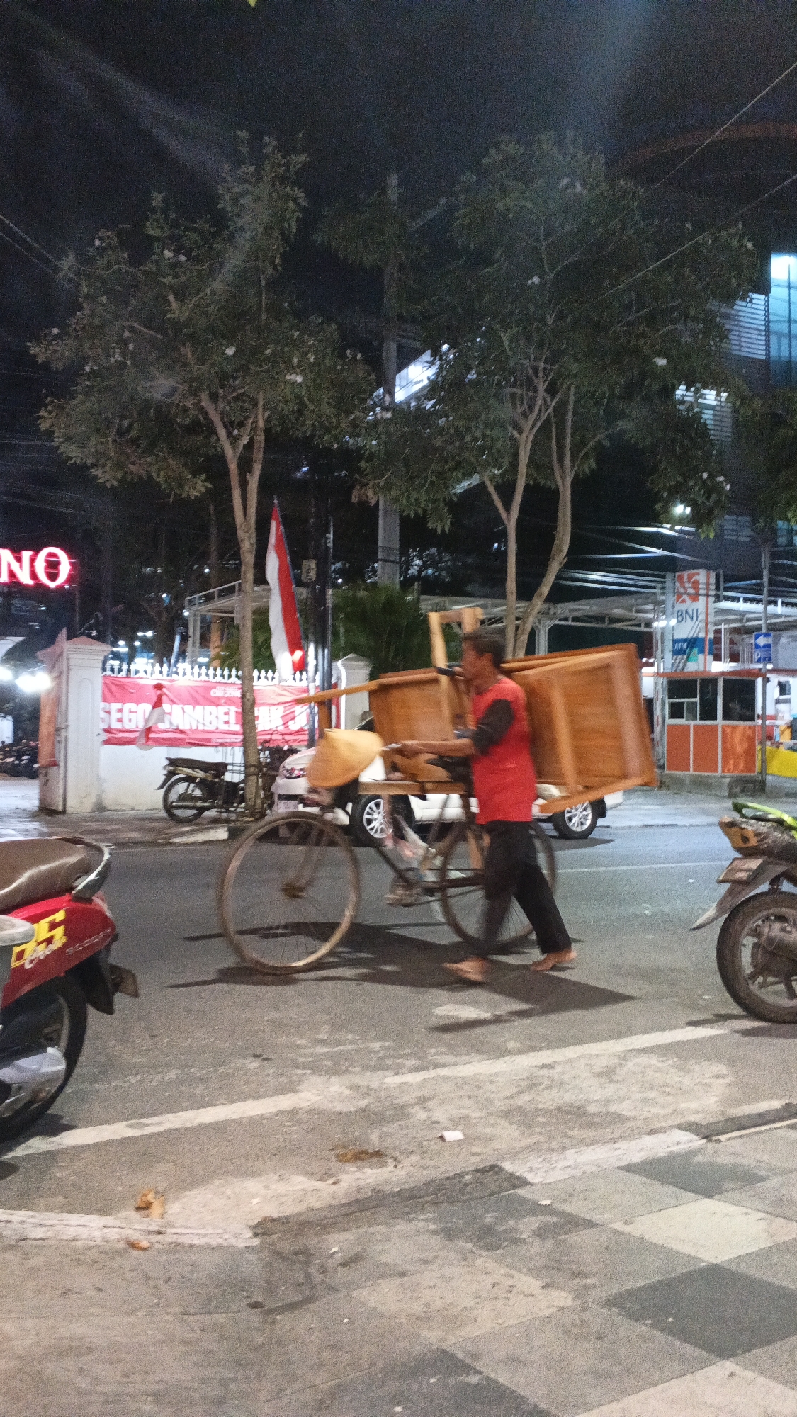 A man is walking on a street at night in Bojonegoro, Indonesia, pushing a cart loaded with wooden furniture. He is wearing a red vest over a dark shirt and black pants, and is barefoot. The cart appears to be a bicycle with an extended frame and two large wooden crates secured to it. Behind the man, a red and white Indonesian flag is visible, as is a sign that reads "SEGO GAMBEL JK". In the background, there are trees, streetlights, buildings, and parked motorcycles. The scene is illuminated by artificial lights, creating shadows and highlights on the wet pavement.