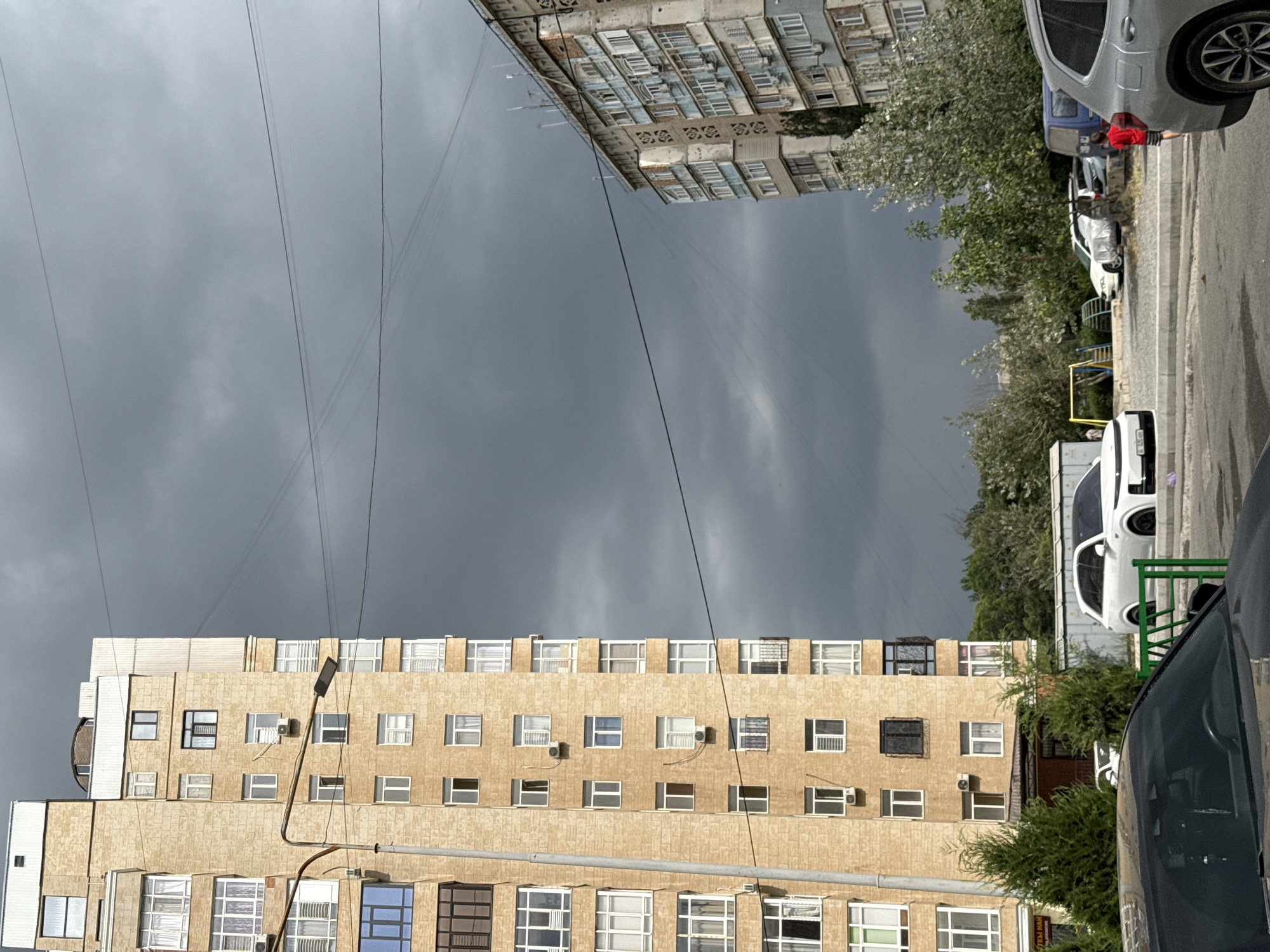 An outdoor, eye-level, medium shot captures a street scene in an urban environment under a dark, stormy sky. The dominant subjects are two tall apartment buildings, one on the left and one on the right of the frame, with the one on the left being closer and more detailed. The left building is a light-colored brick structure with numerous windows, some of which are partially visible and appear to have balconies. A streetlamp with a curved arm extends from the left side of the building towards the street.
The building on the right is older and appears to be constructed of concrete with lighter colored panels and balconies. It is partially obscured by trees and appears to be set further back.
In the foreground, the hood and windshield of a black car are visible. To the right of the black car is a silver SUV. Further back, a white SUV is parked. A small playground with yellow and blue structures is visible behind the white SUV, along with a blue van. A person in a red shirt and striped shorts is standing near the blue van.
The sky is filled with dark, ominous clouds, suggesting impending rain. Power lines are visible crisscrossing the sky. Trees and greenery are scattered throughout the scene, providing some contrast to the urban structures. The overall atmosphere is somewhat somber due to the weather conditions.