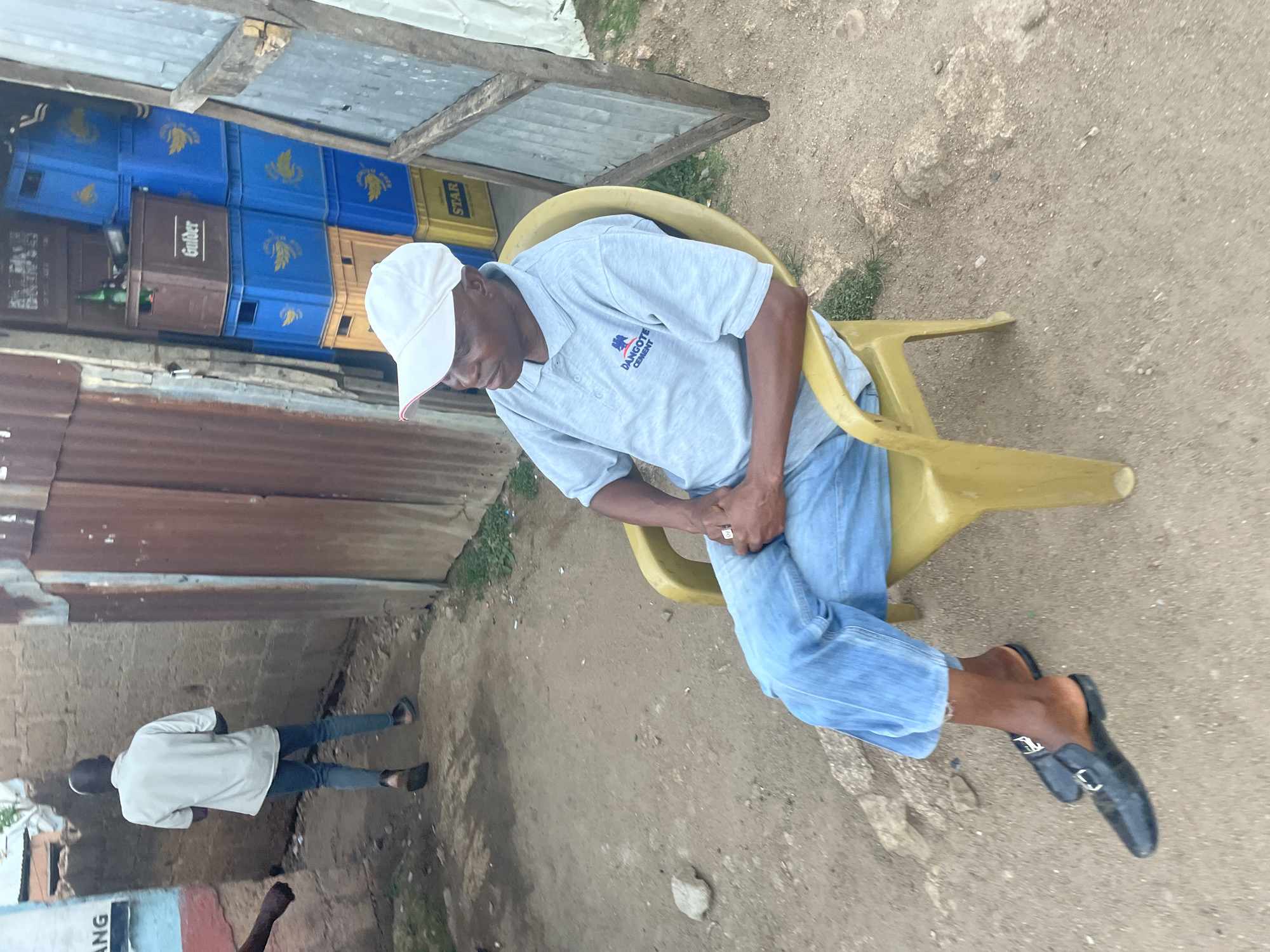 The scene shows a man seated on a yellow plastic chair. He is wearing a light gray short-sleeved shirt with the logo "DANGOTE CEMENT," light blue jeans, a white cap with a red brim, and black loafers. He is looking down at his hands, which are clasped together. A stack of beverage crates is visible in the background, including brands like "STAR" and "Gulder." Another person wearing a white shirt and jeans is walking away. The ground is a sandy, dirt-like surface. The location appears to be an outdoor space in Jos, Nigeria.