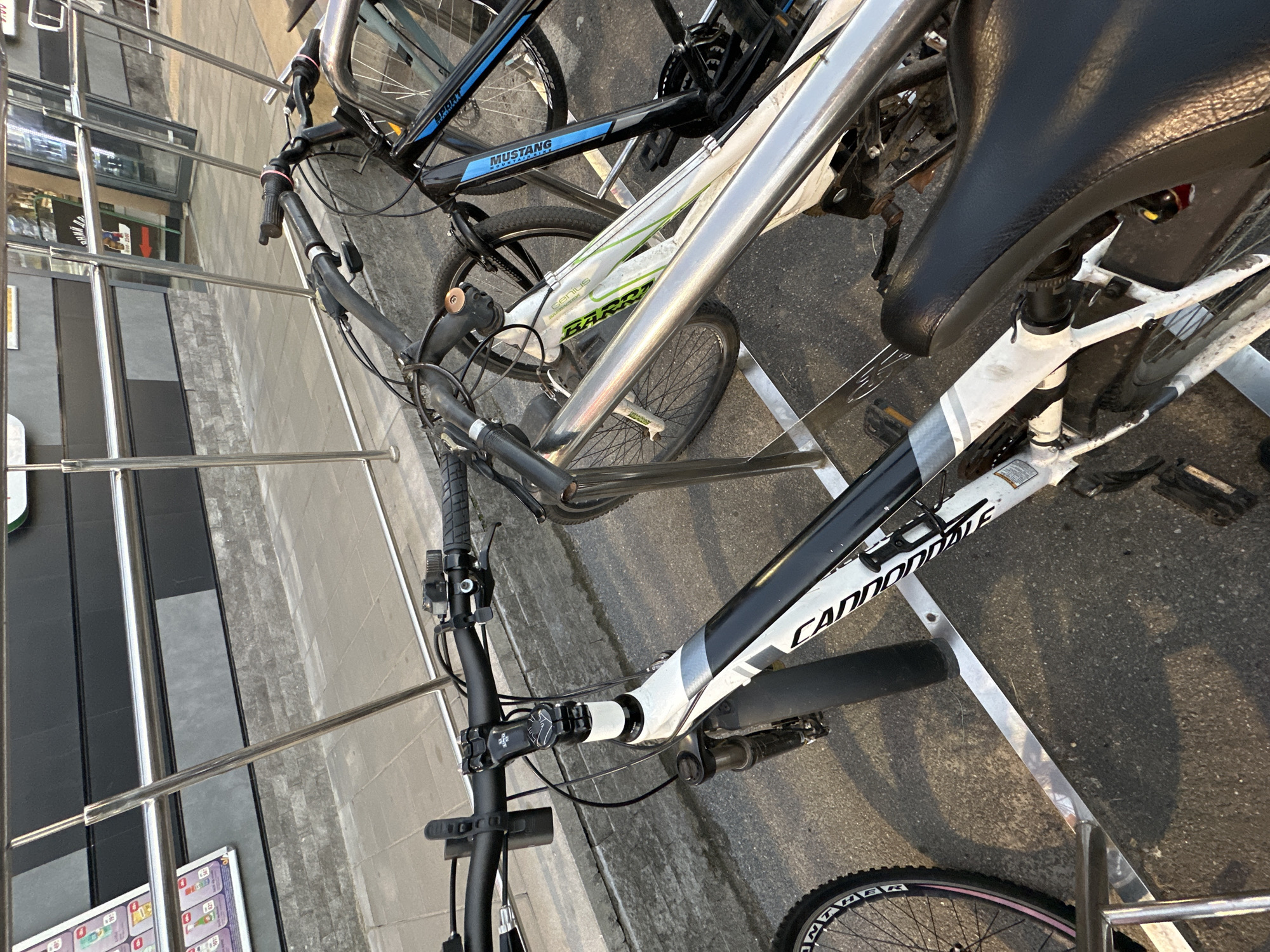 In the city of Unknown, Unknown, several bicycles are parked at an outdoor bike rack. The foreground features a white and black mountain bike, with its handlebars and front fork clearly visible. To the right, a black bicycle with blue accents is partially visible, and another bicycle with a green frame is also present. The bike rack is made of metal, and behind it, a grey stone wall and a sidewalk are visible, leading to a building with reflective windows and black paneling. The scene is illuminated by natural light, casting shadows on the ground.