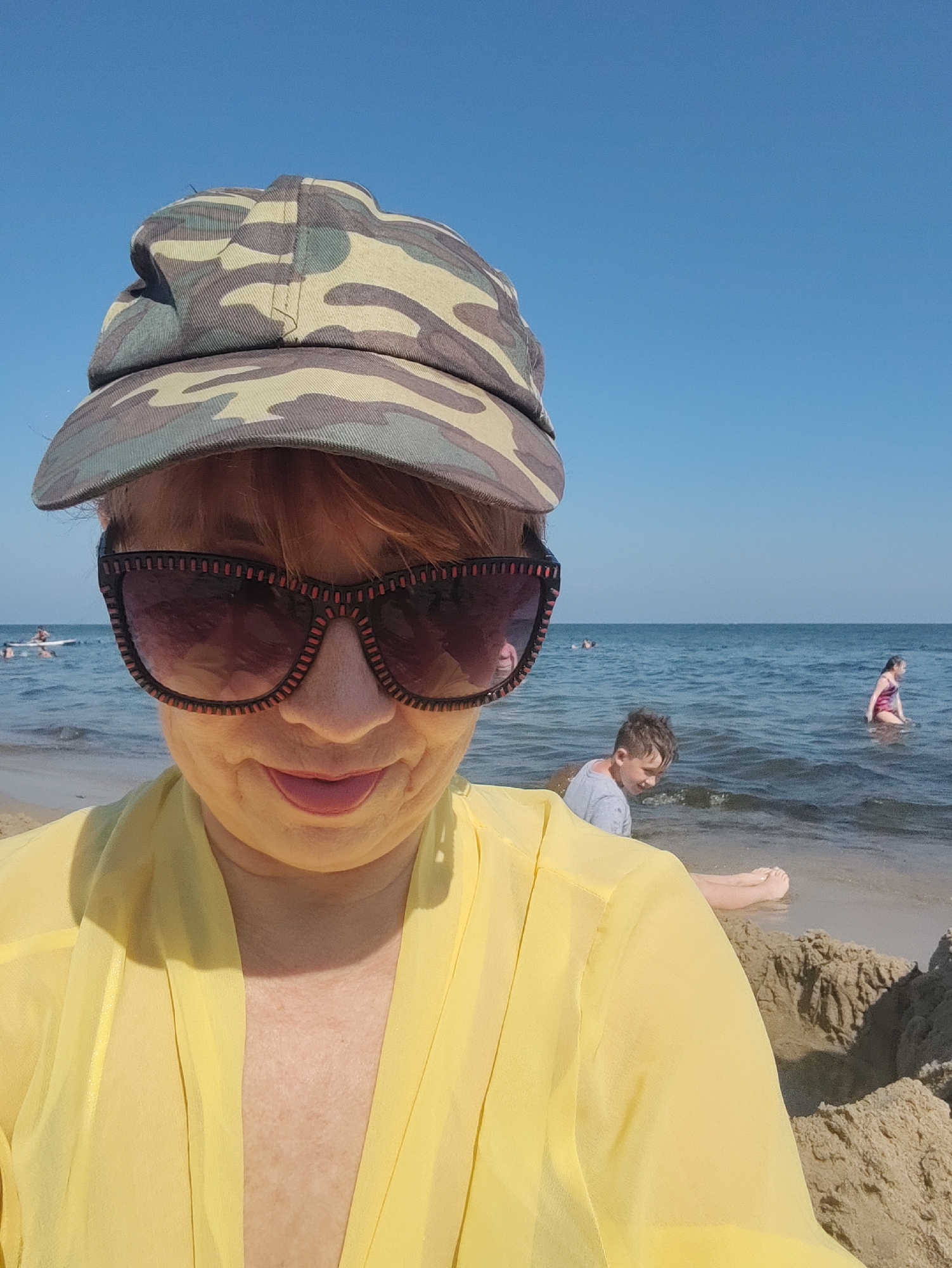 A woman wearing a camouflage cap and sunglasses takes a selfie on a sunny day at the beach in Pionersky, Russia. She is wearing a yellow cover-up over her shoulders. In the background, the sea is calm, and a few people are visible in the water. One person, a child, is partially visible in the foreground near the water's edge, facing away from the camera. The sky is clear and blue, with no clouds. The sand is light brown.