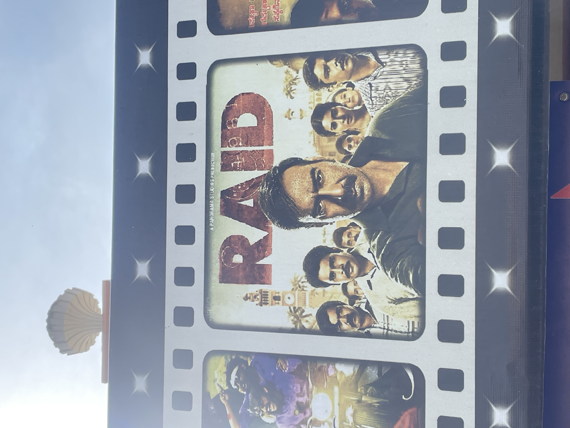 The image shows a poster in Surmaiguda, India. The poster features the Bollywood film "Raid," showing a filmstrip design with images from the movie. A decorative, shell-shaped object is attached to the side of the poster frame. The background includes a bright sky and a blue structure.