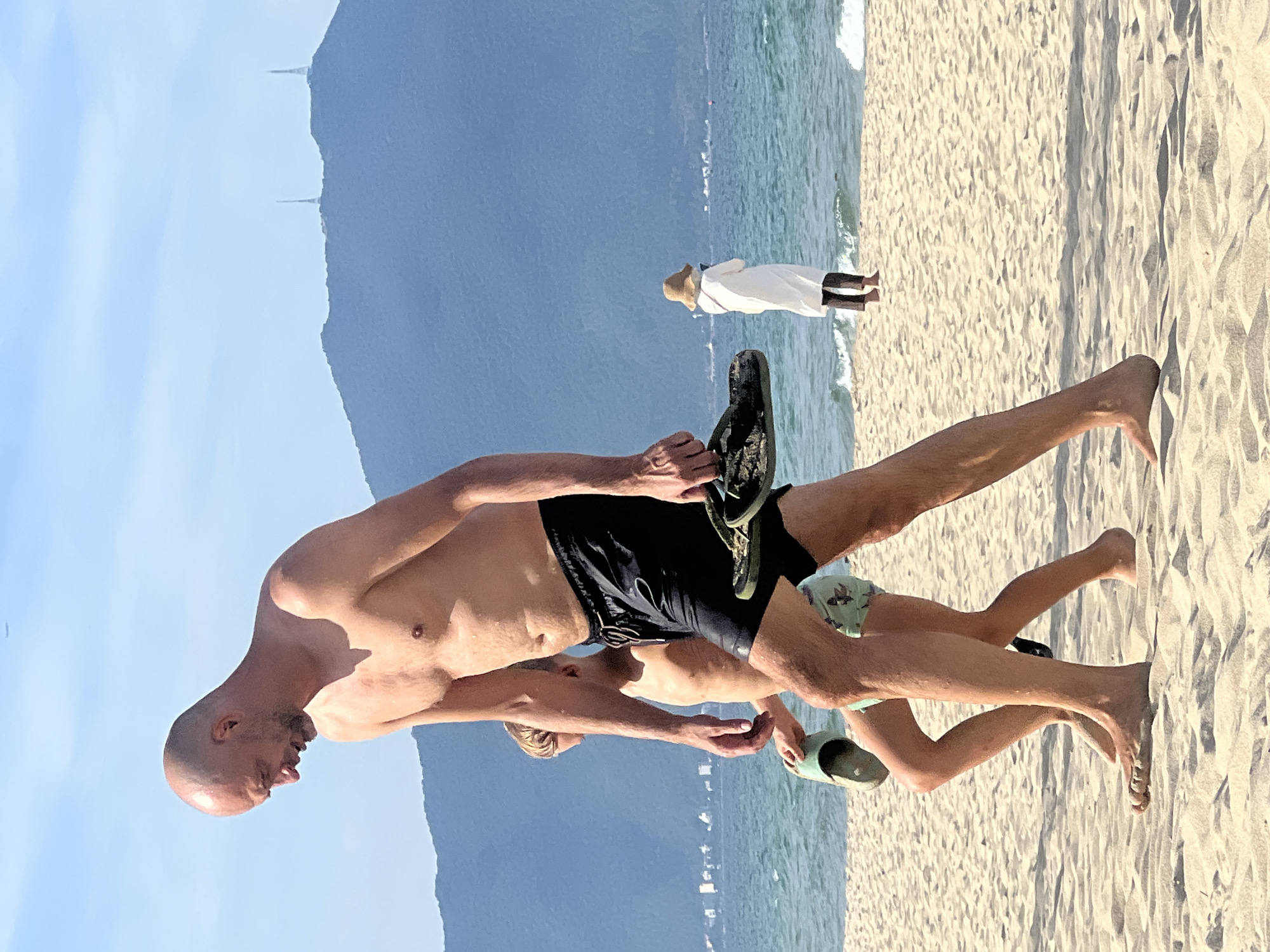 In Đà Nẵng, Vietnam, a bald man in black swim shorts and a young boy in patterned swim shorts walk on a sandy beach. The man carries a pair of dark sandals in his right hand. The boy carries a single light-colored sandal in his left hand. The ocean is visible behind them, with a distant mountain range capped by two towers. In the background, a person in a white robe and a straw hat stands near the water. The sky is clear and blue.