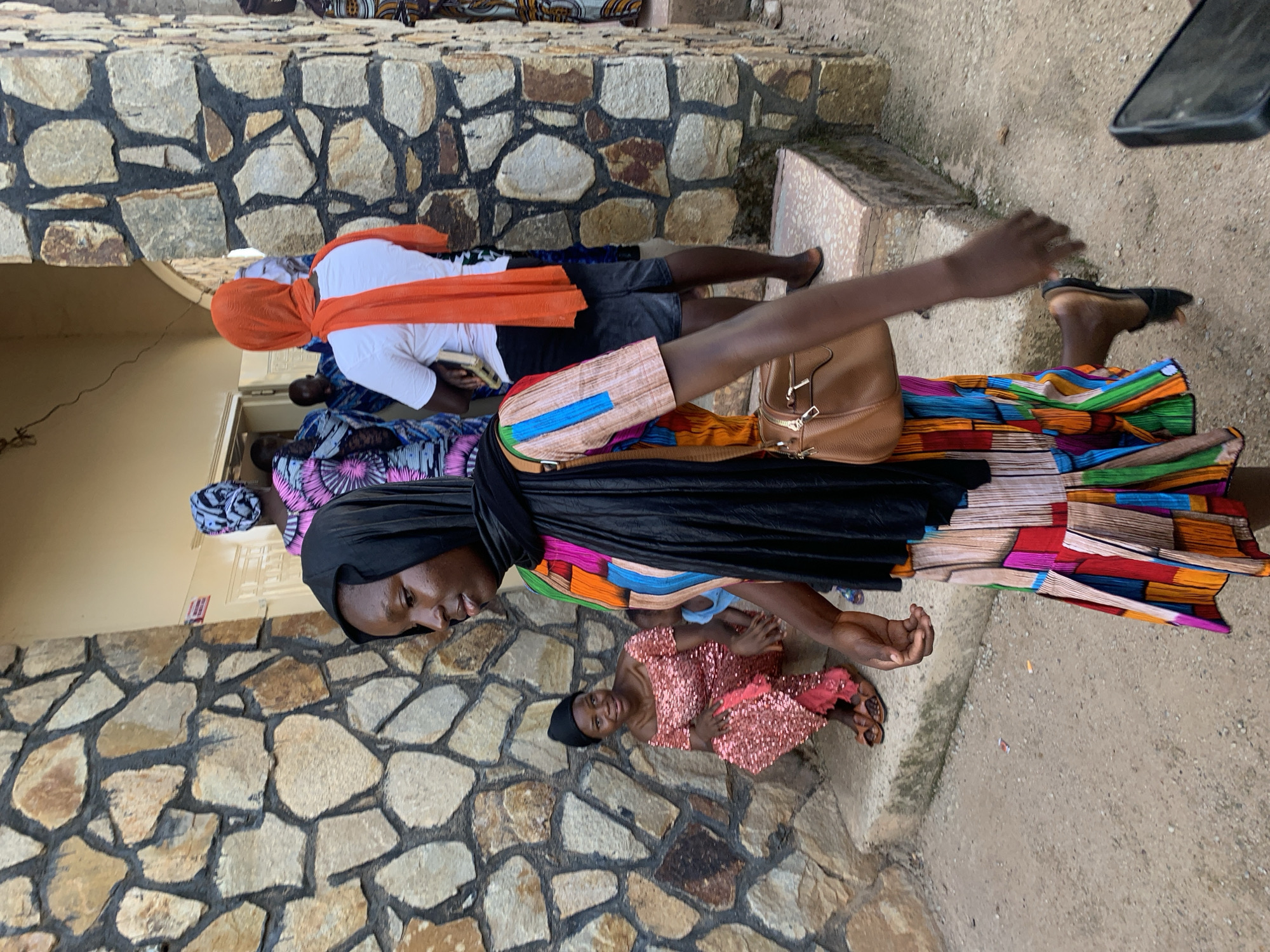 A daytime outdoor scene shows a group of people in Jos, Nigeria. A woman in the foreground is wearing a long, colorful dress with black head covering. A child in a pink sequined dress is next to her. Behind them, other people are also present. One is wearing a white shirt and orange head covering. A brown bag is visible on the side. The background features a stone wall. The setting appears to be a street or public area.