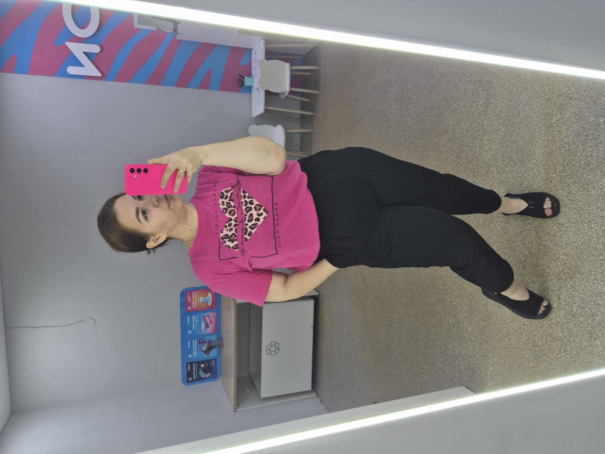 A full-length, mirrored selfie is taken by a woman standing in what appears to be a fitting room or changing area. She is wearing a bright pink t-shirt with a leopard-print heart graphic and black, loose-fitting trousers. Her right hand is in her pocket, and she is holding a bright pink smartphone in her left hand, which is angled to capture her reflection. She has her hair pulled back and is looking towards the camera.
The environment includes a grey wall, a large white bin with a recycling symbol, and a small table with two white chairs in the background. To the right, a tall, vertical light fixture runs along the edge of the mirror. The floor is a speckled grey material. A colorful patterned wall with the letters "ион" is visible in the background on the right side, which is a partial reflection of a logo. Based on the presence of the logo "ион" and the context provided by the user, the location is likely the Ozon fitting room in Chaikovsky, Russia. Ozon is a large Russian e-commerce company.