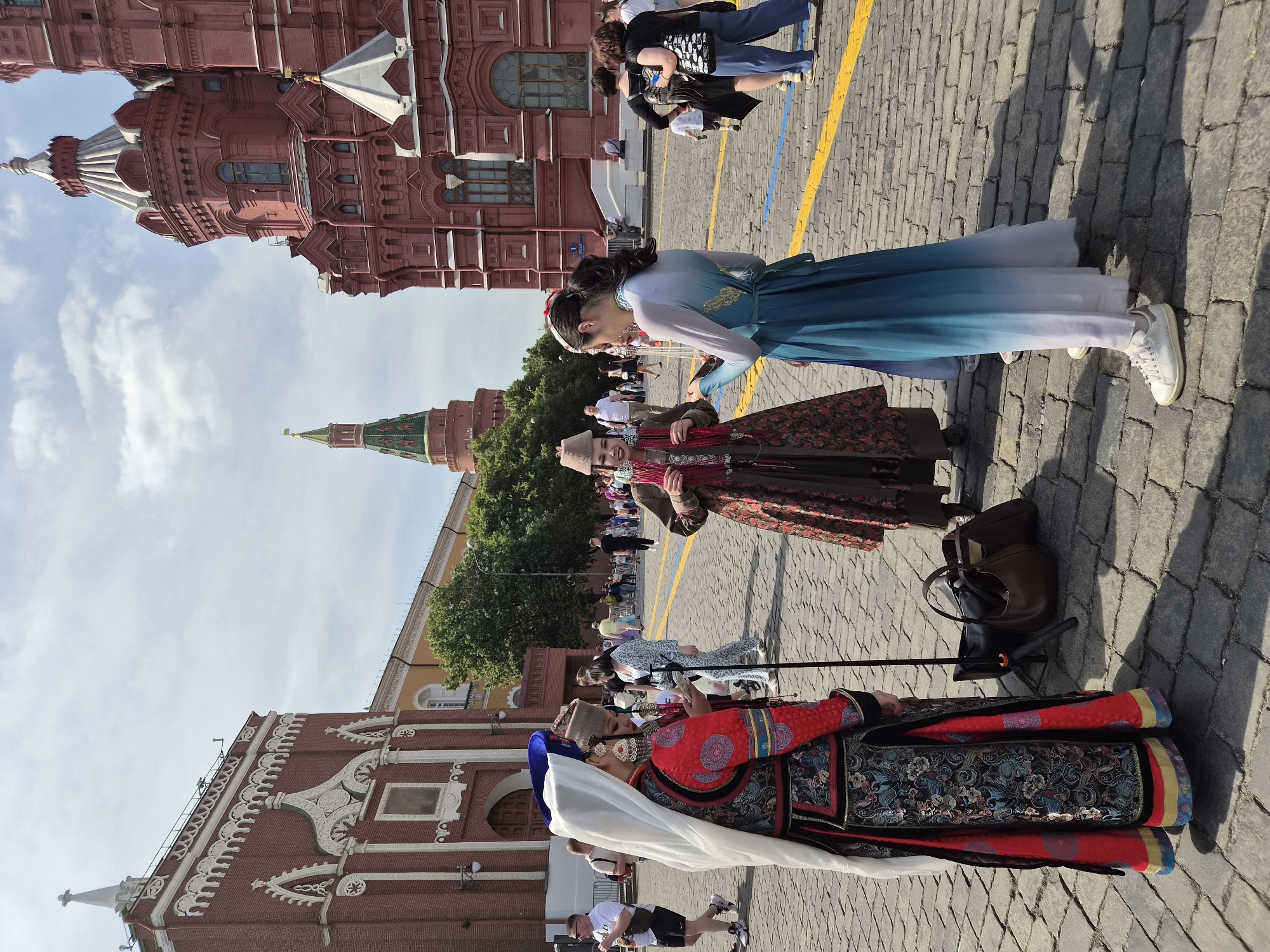 This image depicts a daytime scene on Red Square in Moscow, Russia. In the foreground, three women are prominently featured, dressed in traditional or historically-inspired garments. The woman on the far left, viewed from behind, wears a long, richly embroidered red and black dress with blue and gold accents, a white veil draped over a blue headpiece, and large ornate earrings; a thin black pole rests vertically next to her, with a black and a brown bag on the cobblestones at her feet. The central woman, seen from a three-quarter angle, wears a long, dark patterned coat, a decorative cap, and multiple strands of red adornment around her neck; she faces the woman on the right. The woman on the right, also seen from behind, wears a flowing blue ombre dress that fades to white at the hem, with a subtle gold pattern on the back, and white sneakers; she is turned towards the central woman, appearing to engage with her.
The ground is paved with large, irregular cobblestones, marked with parallel yellow and blue painted lines. In the background, numerous people in casual modern clothing walk across the square. Architectural elements include the ornate red-brick State Historical Museum with its multiple turrets and white detailing on the right, and a section of the red-brick GUM building with white decorative elements on the left. A green-domed tower, identified as the Nikolskaya Tower of the Moscow Kremlin, is visible in the distance between the two prominent buildings. The sky is partly cloudy, and strong sunlight casts distinct shadows on the ground.