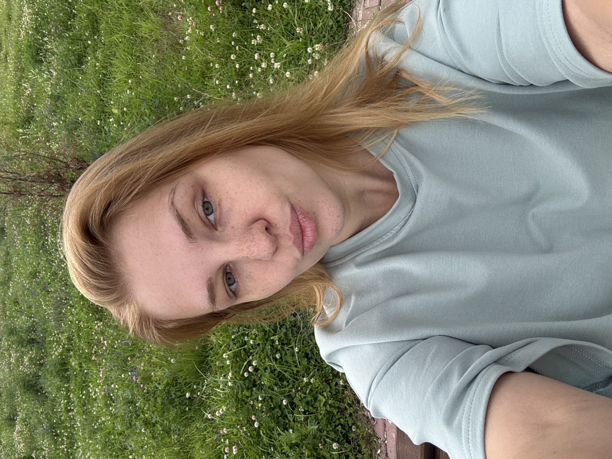 The image is a selfie of a young woman with fair skin, blonde hair, and blue-green eyes. She has freckles across her nose and cheeks. Her lips are slightly parted, and she is looking directly at the camera with a neutral expression. She is wearing a light blue, long-sleeved t-shirt.
The background is a grassy area with small white and pink wildflowers, suggesting a meadow or park. The lighting indicates it is daytime, likely with overcast or soft lighting given the absence of harsh shadows. The scene has a natural, outdoor feel, possibly in a rural or park setting.
The orientation of the image is rotated, with the woman appearing to be lying down or leaning back. The overall impression is of a relaxed, casual moment captured outdoors. There is no visible text in the image.