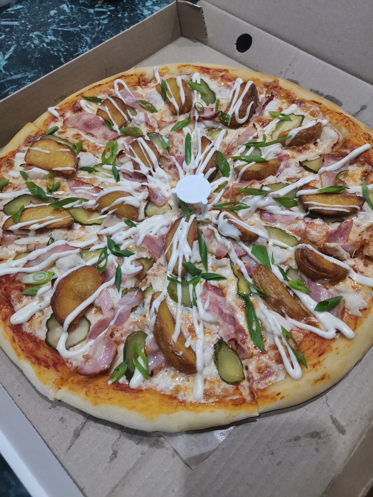 A whole pizza is presented in a cardboard pizza box. The pizza has a thin crust and is topped with cheese, tomato sauce, bacon, sliced pickles, potato wedges, and chopped green onions. A white sauce is drizzled in a pattern over the toppings. The pizza box is open and placed on a dark, speckled surface. The image was taken indoors, likely in a home or restaurant setting in Kizel, Russia, based on the context provided by the user. No people are visible in the image.