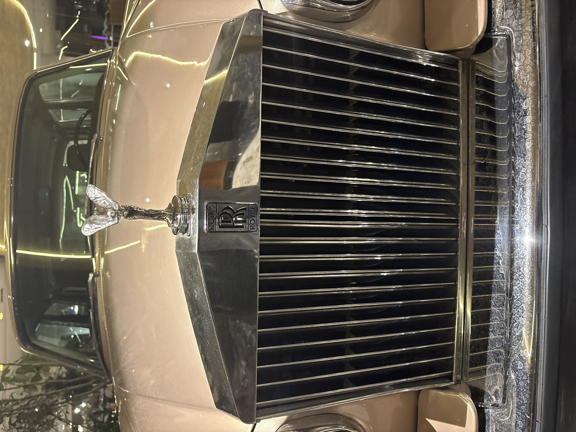 The image shows the front of a Rolls Royce automobile. The car is tan with a silver grill. The Spirit of Ecstasy emblem is visible on the hood. The location is potentially in Mexico City, Mexico.