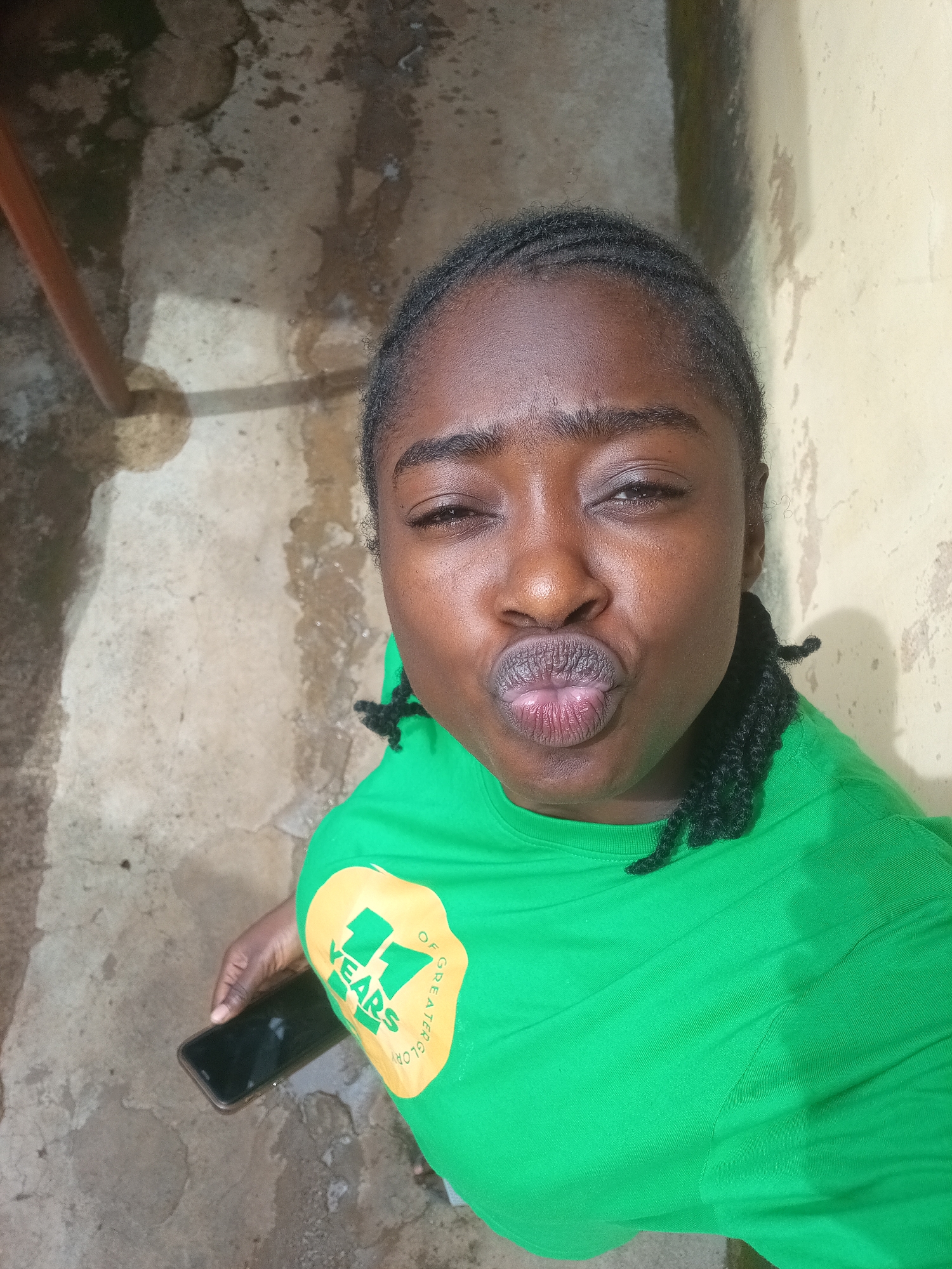 A person with dark skin and braided hair is taking a selfie. They are wearing a bright green t-shirt with a yellow circular logo on the left side of the chest that says "11 YEARS OF GREATER GLORY". The person is making a kissing face. They are holding a black smartphone in their left hand. The background appears to be an outdoor, urban setting with a textured concrete ground and a light-colored wall. There is a thin brown pole on the left side of the image. The lighting suggests it is daytime. The location is Unknown, Unknown.