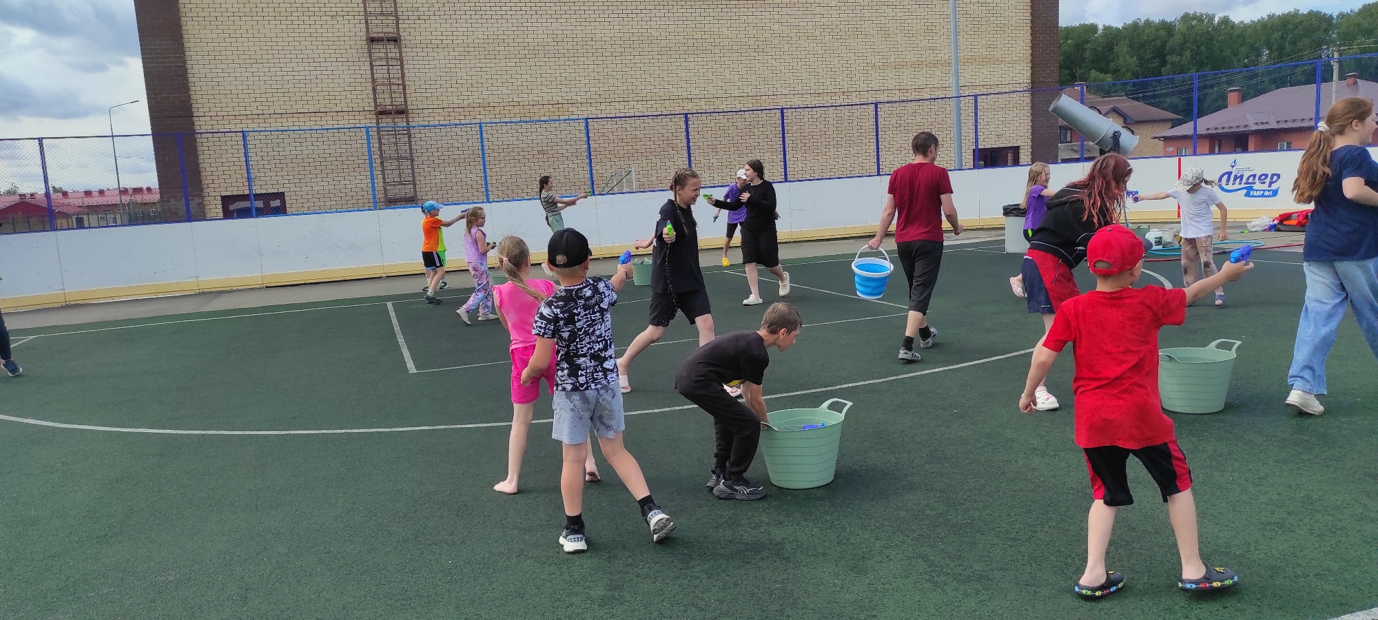 The image depicts a group of children and adults engaged in a water gun fight on a green artificial turf field, likely a sports court, located in Noviy, Russia. The background features a brick building with a blue fence and a ladder. Several children are actively participating, holding water guns and buckets. One boy in the foreground, wearing a red cap and t-shirt, is aiming a blue water gun. Another child, in a black t-shirt, is bent over a large green bucket. Adults are also present, with one woman in a red shirt carrying a blue bucket. The overall atmosphere suggests a recreational event or party.