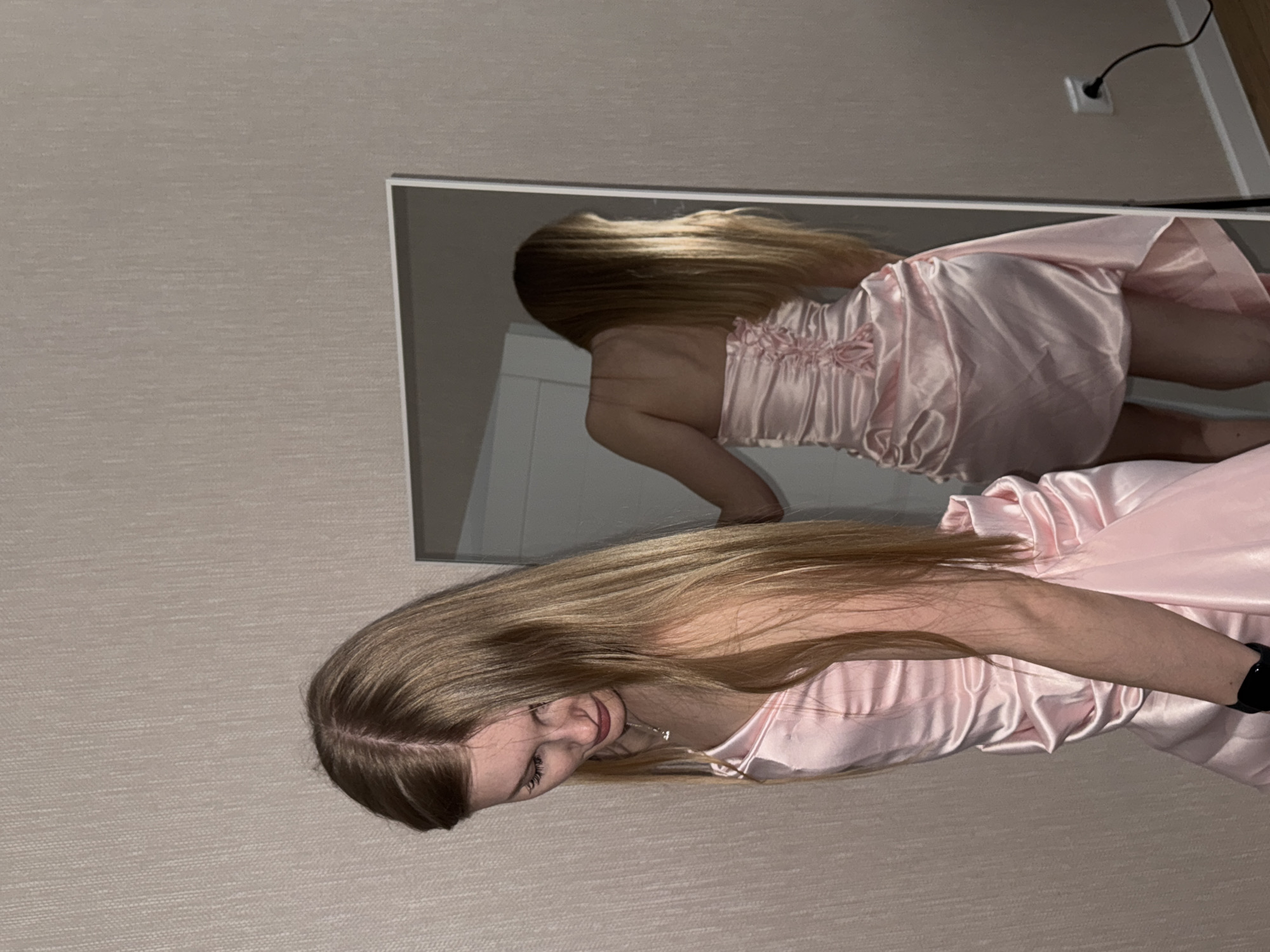 A woman is standing indoors in front of a mirror. The woman has blonde hair and is wearing a pink dress. The location is presumed to be in the city of Perm, Russia, though no specific identifiable landmarks are present.