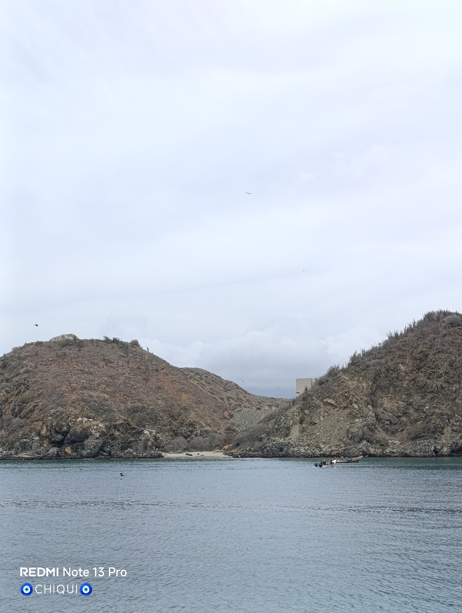The image depicts a seascape, likely a bay or cove, under a cloudy sky. Two hills or small mountains frame the scene. The hills are covered in sparse vegetation and rocky terrain. A building or structure is visible between the hills. In the water, a small boat with several people on board can be seen. The water is calm with slight ripples. The location is likely in Porlamar, Venezuela.