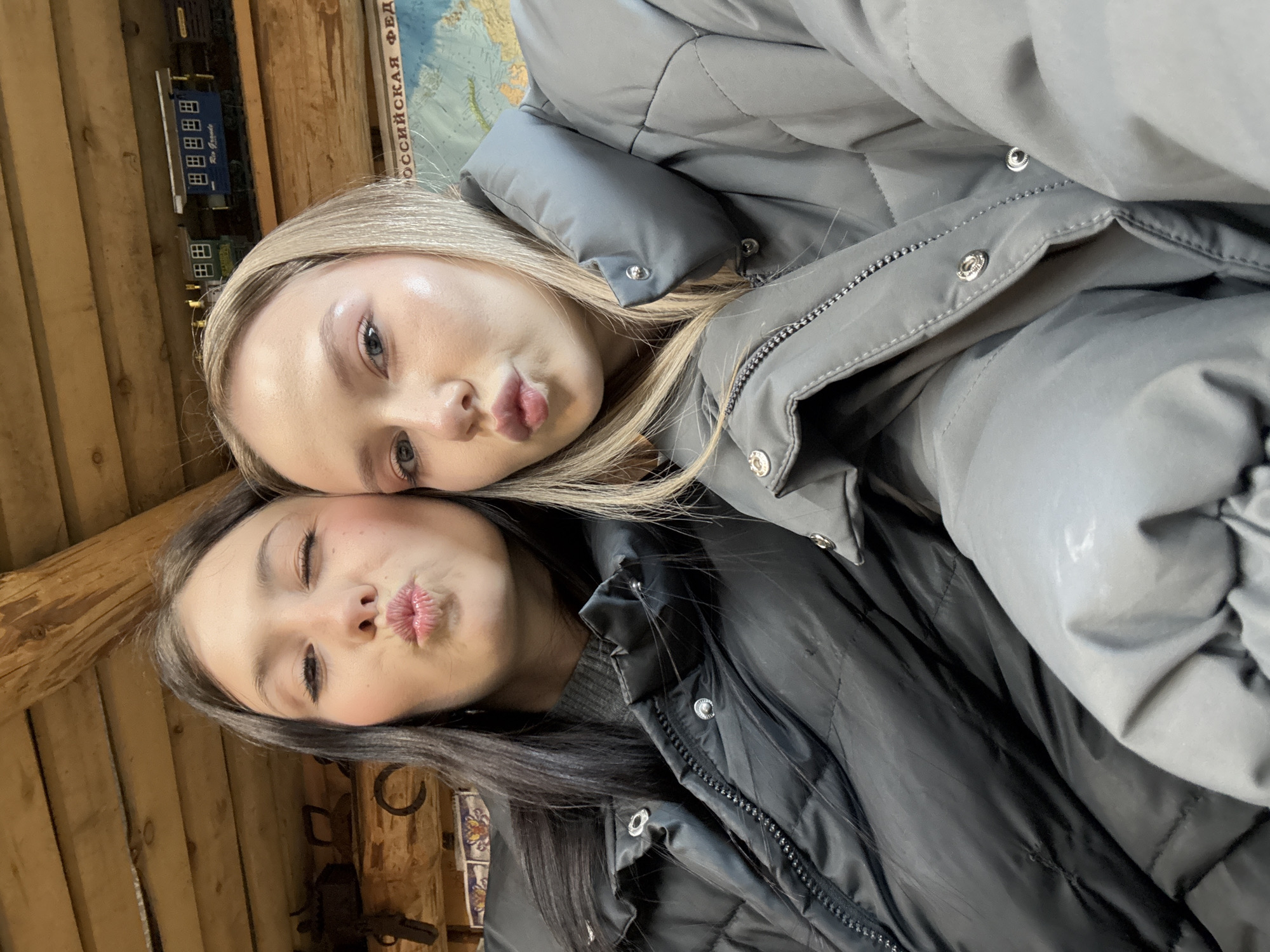 Two young women are posing for a selfie. The woman on top has blonde hair and is wearing a grey puffer jacket. The woman on the bottom has dark hair and is wearing a black puffer jacket. Both women are pouting their lips towards the camera. The background shows a wooden wall and a map of Russia. Based on the Russian lettering on the map, the location might be Фоки, Russia. There are also some decorative items hanging on the wall.