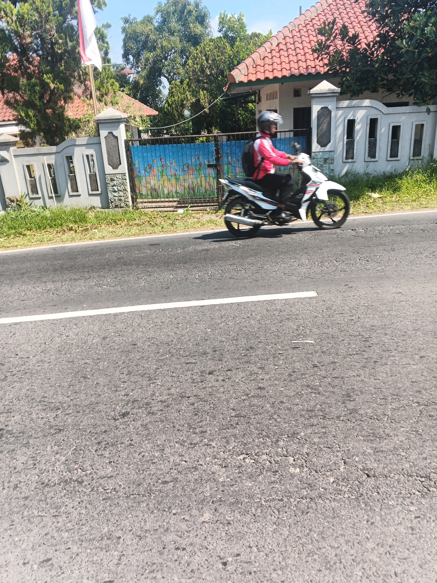 A person wearing a pink jacket and a helmet rides a white motorcycle down a paved road in Bandungan, Indonesia. The motorcycle is moving from left to right across the frame. The road has a white stripe down the center. To the right of the road, there is a grassy area and then a fence with a mural of people dancing. Beyond the fence, there is a house with a red tiled roof and windows. There are trees and a flagpole with a red and white flag in the background. The sky is blue and clear. The image is taken from a slightly elevated perspective.