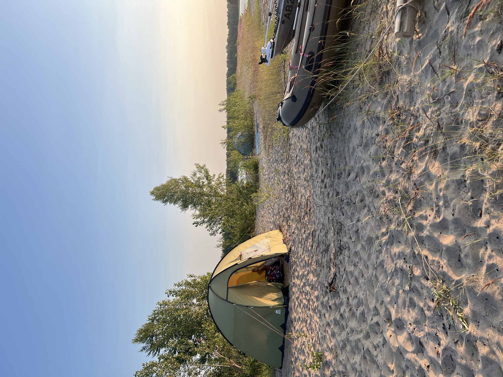 This image depicts a camping scene on a sandy beach, likely near a body of water given the presence of inflatable boats. A green tent is set up on the sand, its entrance open, revealing a glimpse of bedding inside. In the foreground, two inflatable boats are resting on the sand. One boat, positioned closer to the viewer, is a dark green, and another, further back, is a lighter grey with "AB2" visible on its side. In the background, there are trees and vegetation, and a hazy horizon suggests a vast open space, possibly a lake or river, under a clear sky with soft, warm lighting indicative of either sunrise or sunset. The location is identified as Borovoe, Russia. The scene appears peaceful and undisturbed.