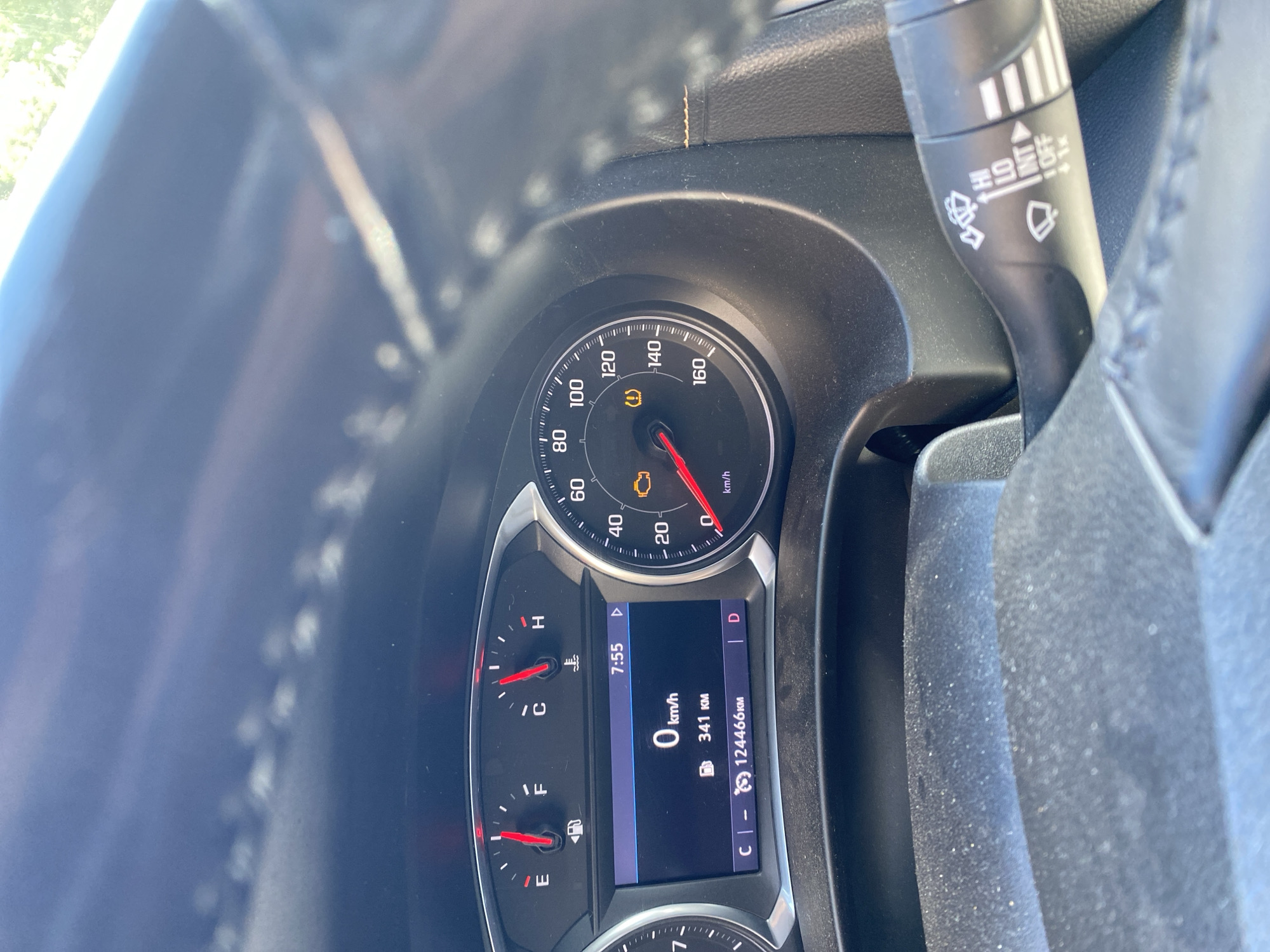 The image shows a close-up view of a car's dashboard. The speedometer is visible, with the needle indicating 0 km/h. The fuel gauge shows the tank is nearly full, and the temperature gauge is in the normal range. A digital display shows the time as 7:55, the trip odometer reading 341 km, and the total odometer reading 124466 km. A warning light shaped like an engine is illuminated, as is a tire pressure warning light. The steering wheel is partially visible, along with a windshield wiper control stalk. The background is blurred, suggesting the photo was taken inside a moving vehicle. The location is not specified.